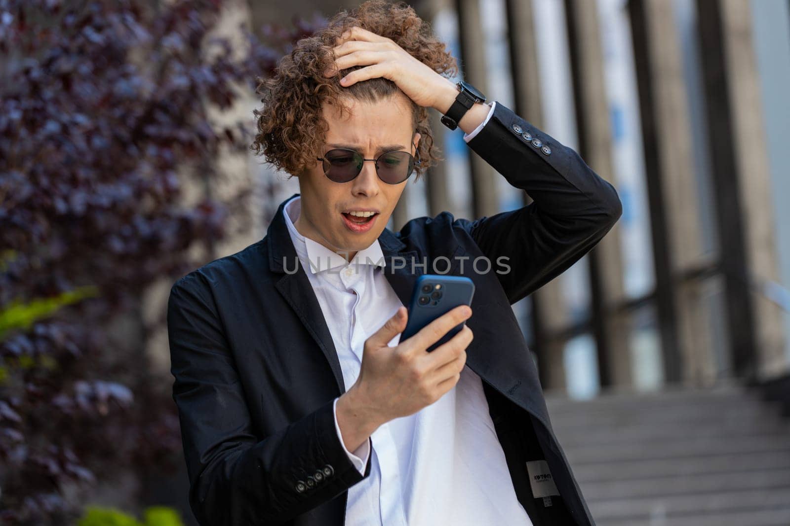 Shocked young businessman using smartphone typing browsing, loses surprised by bad news, fortune loss, fail bankruptcy outdoors. Sad male guy with curly hair in downtown city street. Business people.