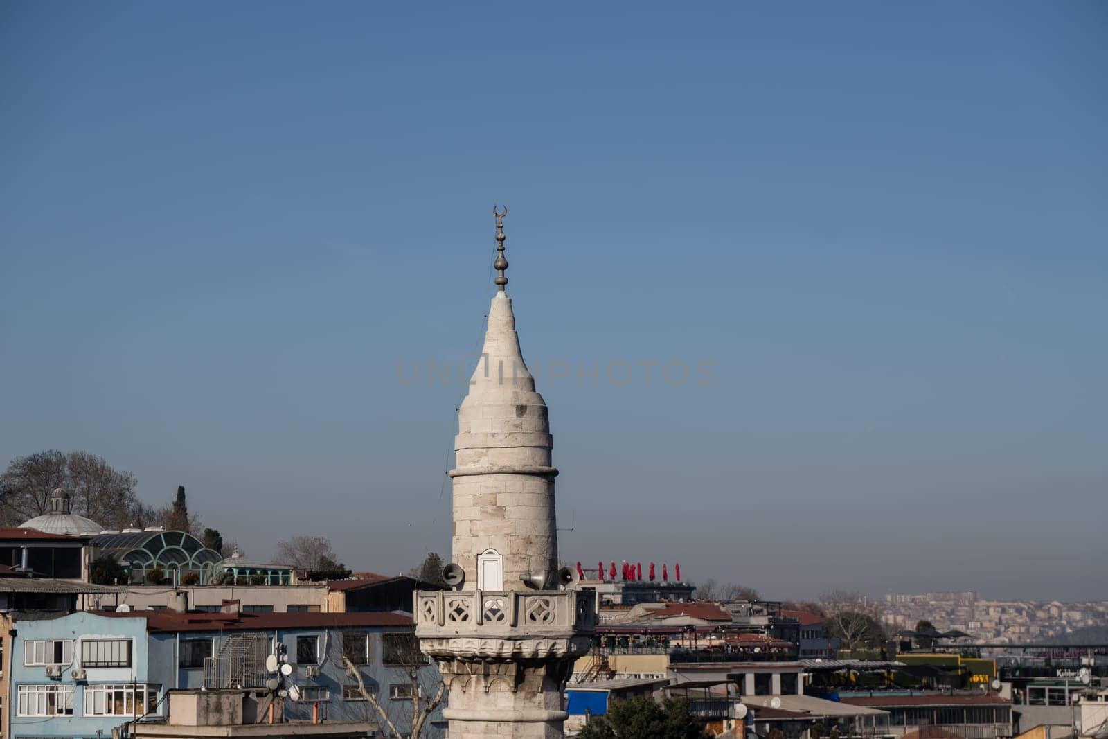 A slender, conical minaret, adorned with ornate detailing, rises above a dense cluster of low-rise buildings under a clear blue sky. The scene suggests a cityscape in a Middle Eastern or Mediterranean setting.
