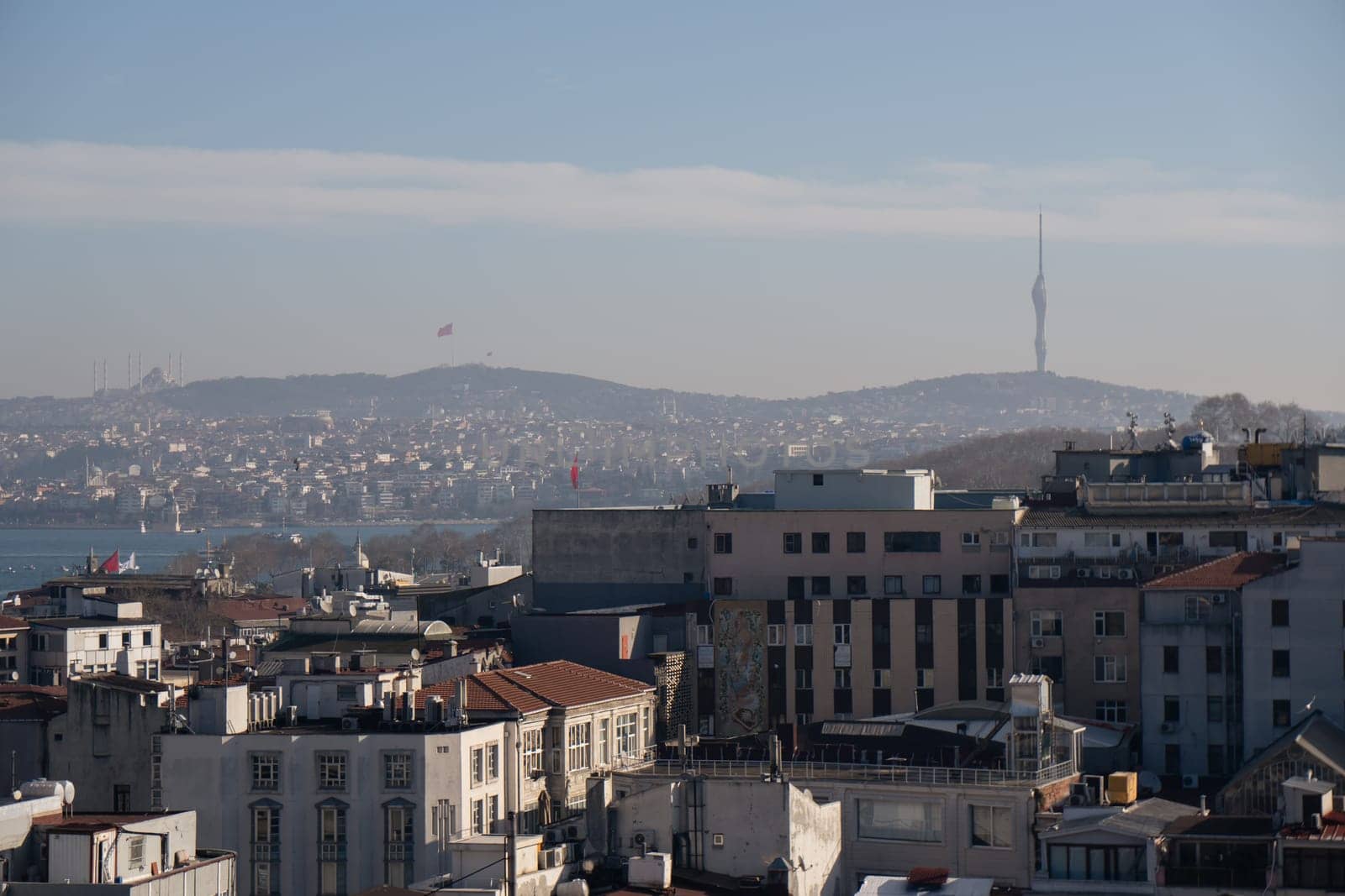Elevated view of Istanbul's cityscape. Densely packed buildings, hills, and a distant communication tower under a clear sky. Water body visible in the background.