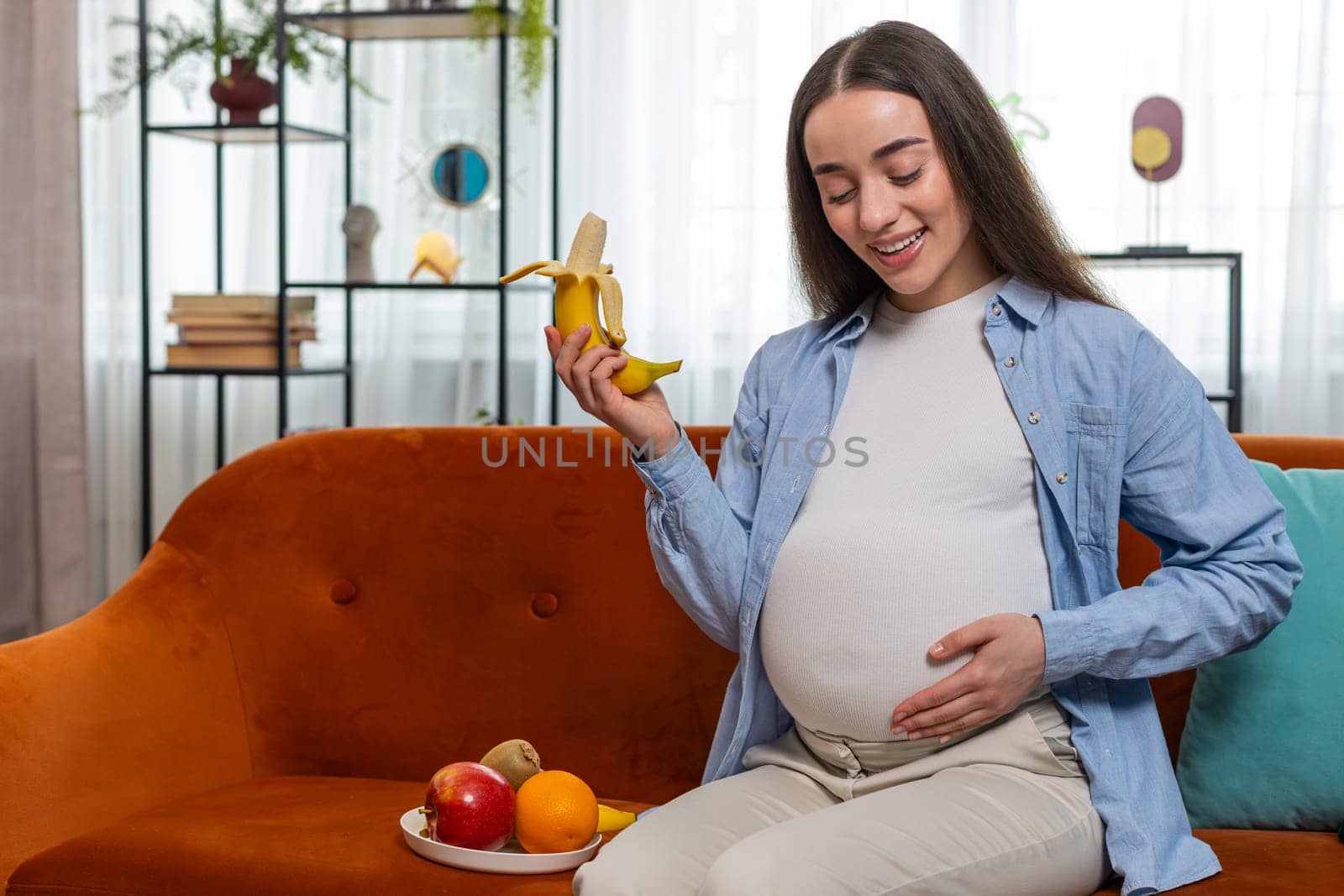 Happy pregnant woman with great appetite peels and eats banana fruit sits on sofa in living room with modern interior. Motherhood self-care childcare. Smiling Caucasian lady chooses right healthy food