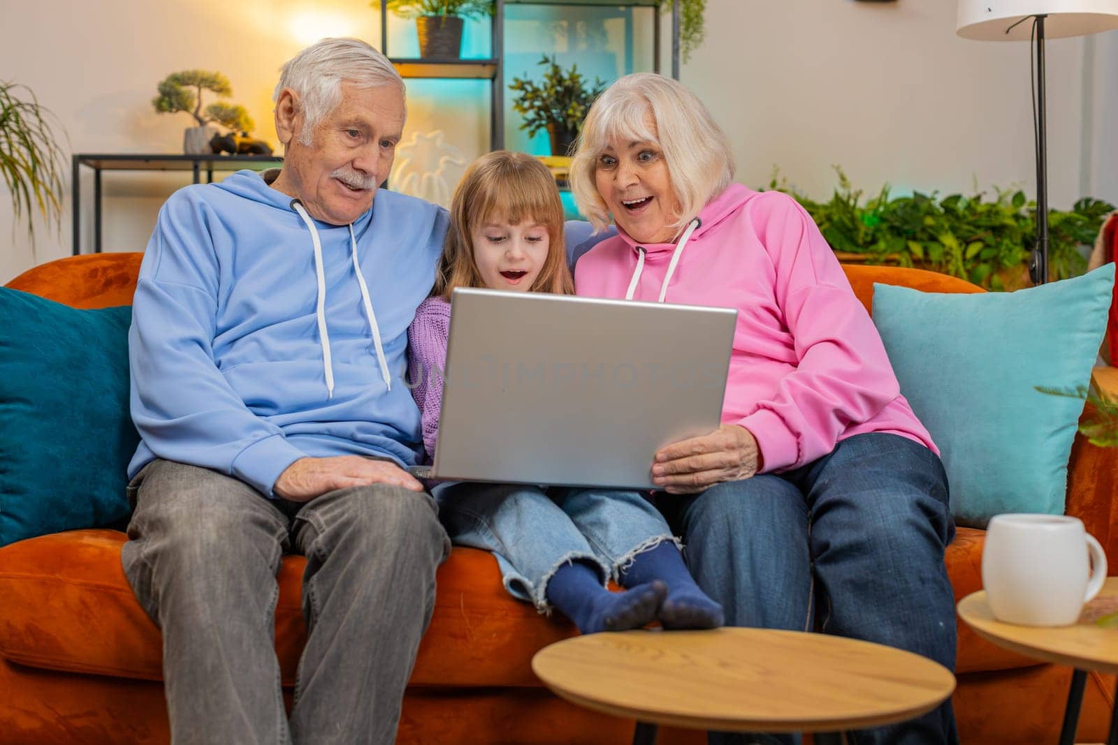 Elderly Caucasian couple man and woman with child girl granddaughter spending time at home together, sitting on sofa in the living room, using laptop computer. Watching videos playing games on weekend