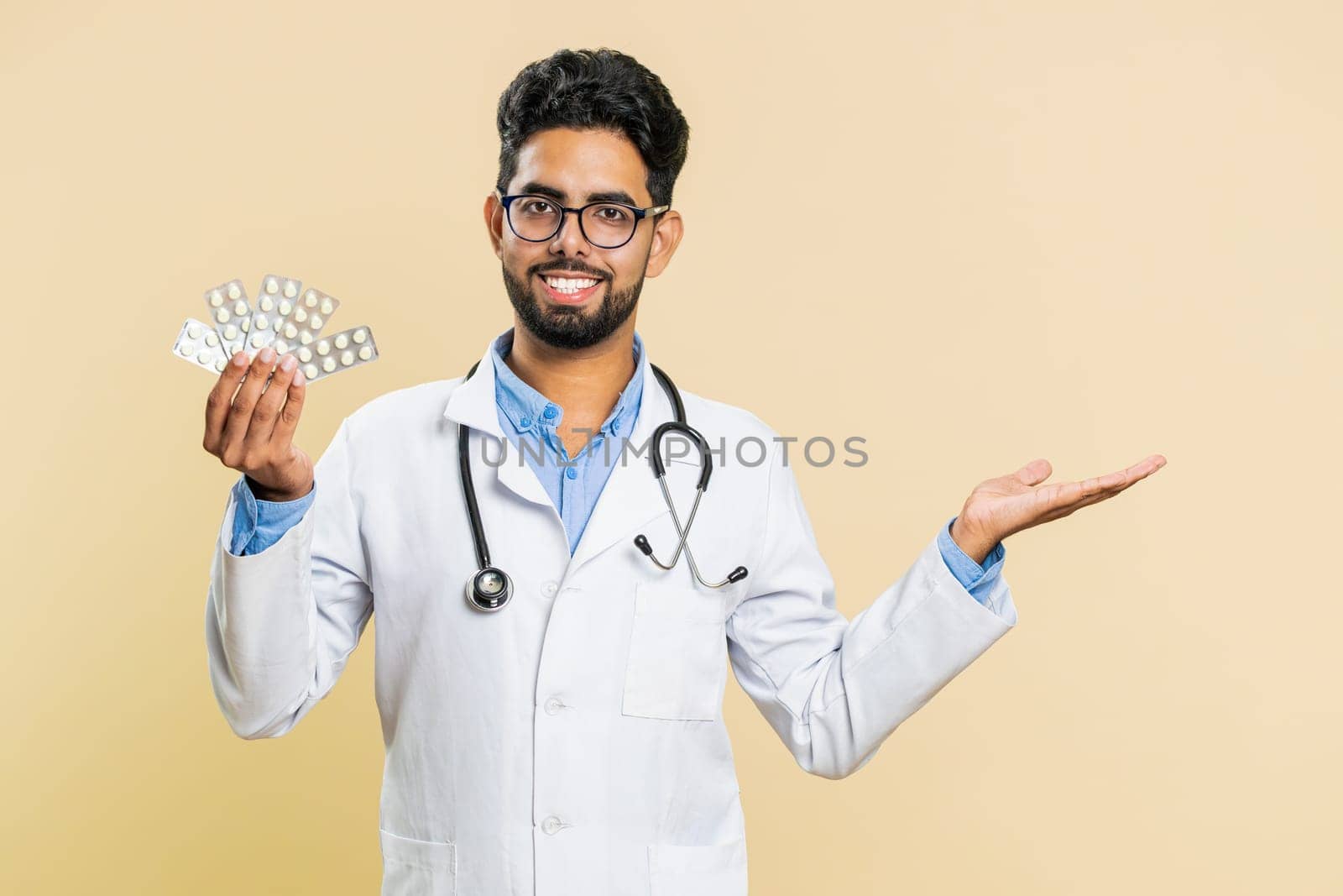 Indian doctor cardiologist man shows lot of tablet pills in blisters, recommends drugs vitamin antibiotic cure treatment. Health care. Arabian apothecary pharmacist guy isolated on beige background
