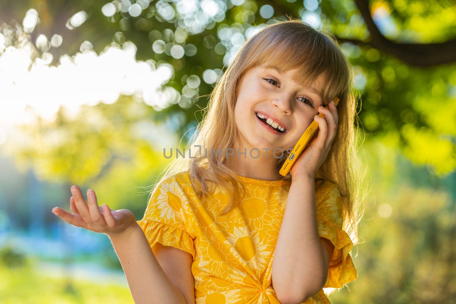 Happy preteen child kid having remote conversation communicate speaking by smartphone. Lovely blonde girl daughter talking with mother on phone good news gossip standing on city sunset park street