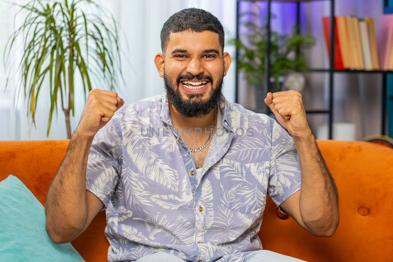 Happy Indian man shouting, celebrating success, winning, goal achievement good news, lottery luck, victory, watching TV championship at modern home apartment. Arabian guy in living room sits on couch