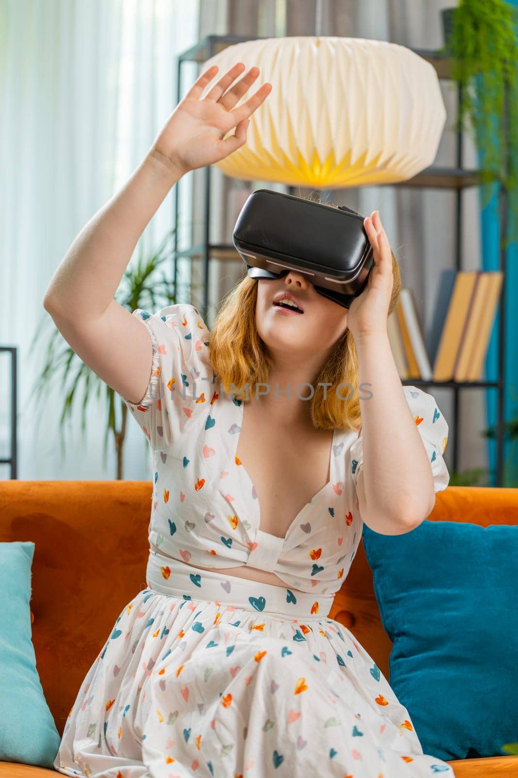 Woman using virtual reality futuristic technology VR app headset helmet to play simulation 3D 360 video game, watching film movie at modern home apartment. Girl in goggles sitting on couch. Vertical