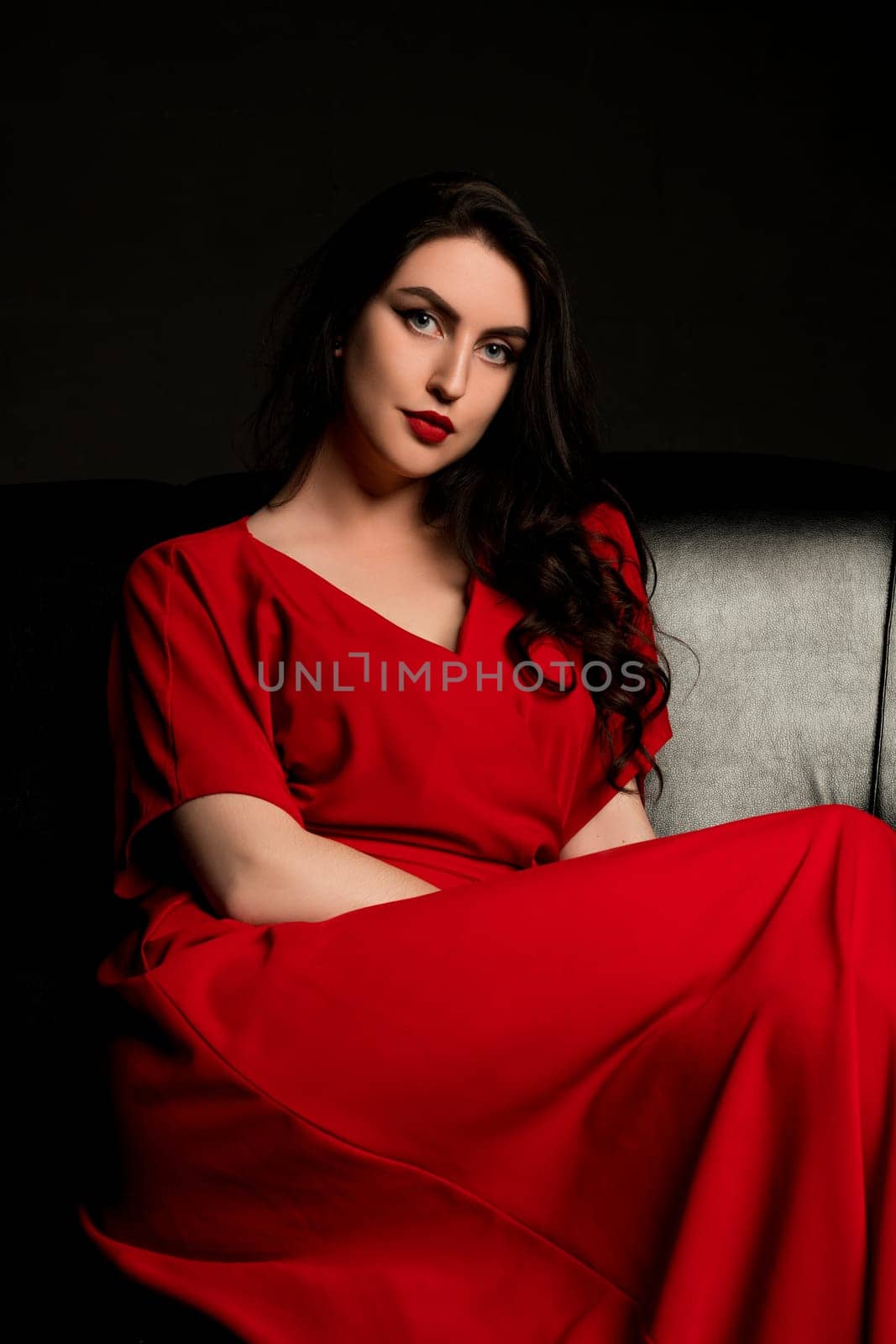Sexy brunette in red dress sitting on black leather sofa shot in the dark room