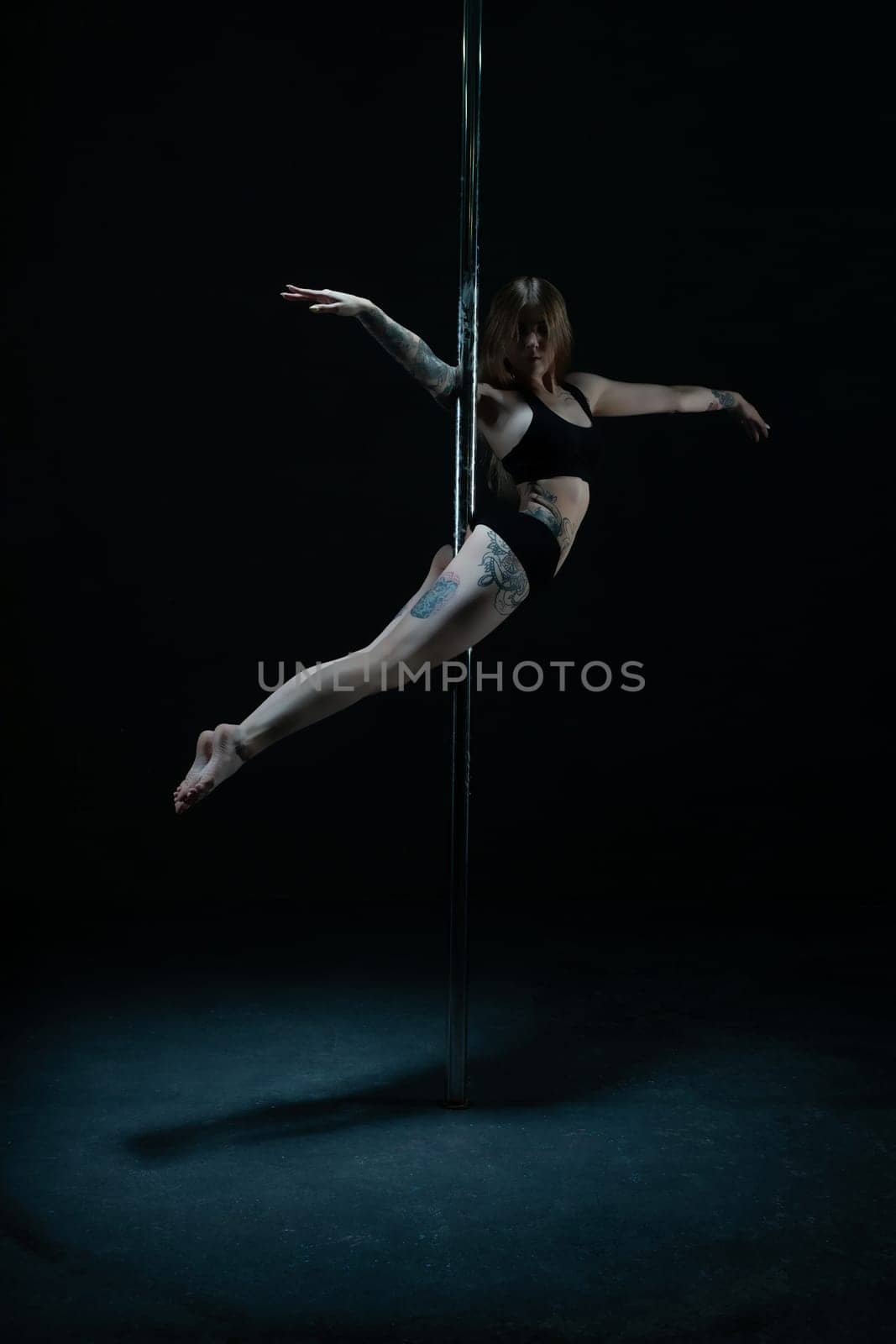 Young flexible woman with tattoos posing on pylon with outstretched arms on black background