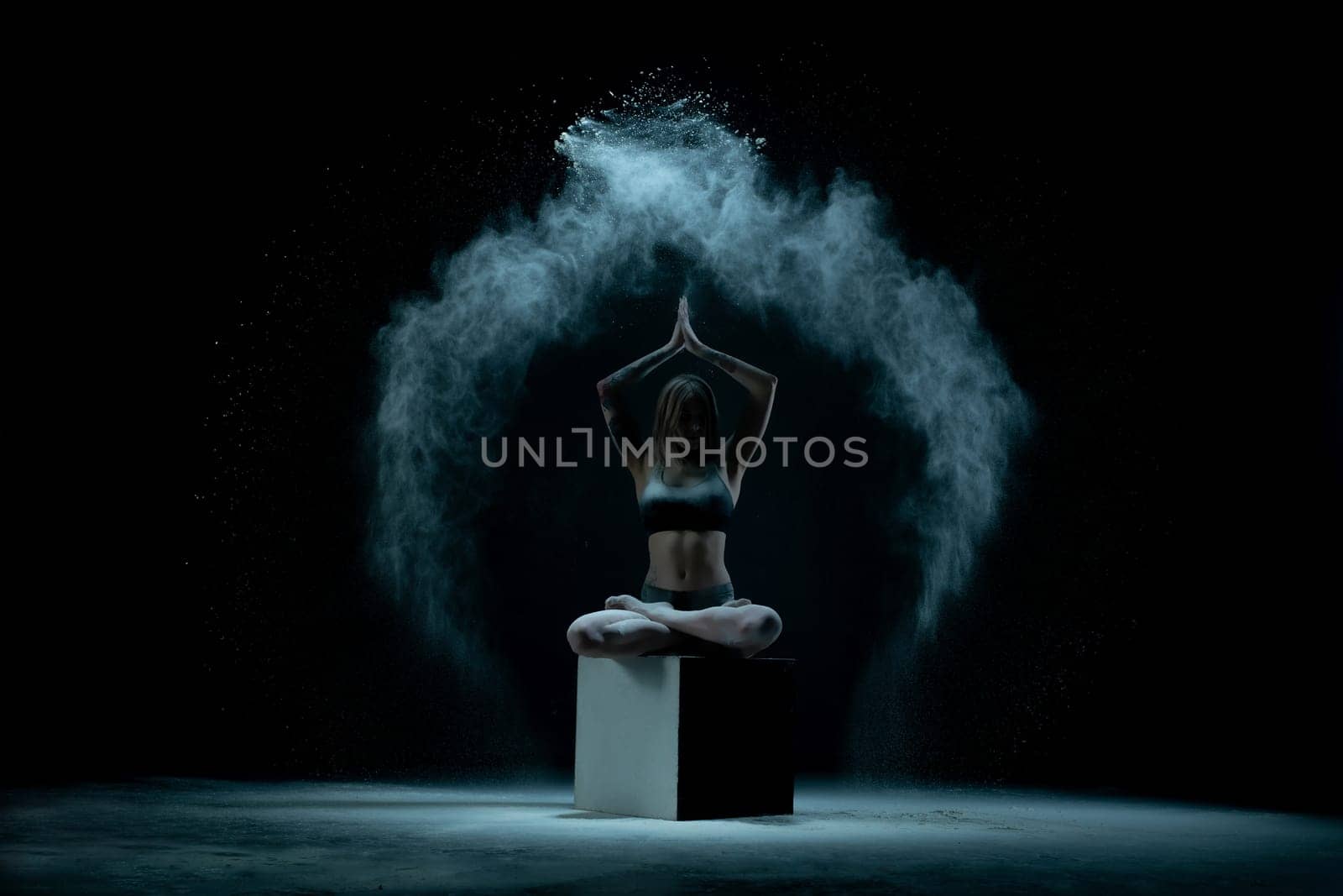 Young flexible woman in lingerie posing on cube while performing in cloud of white dust