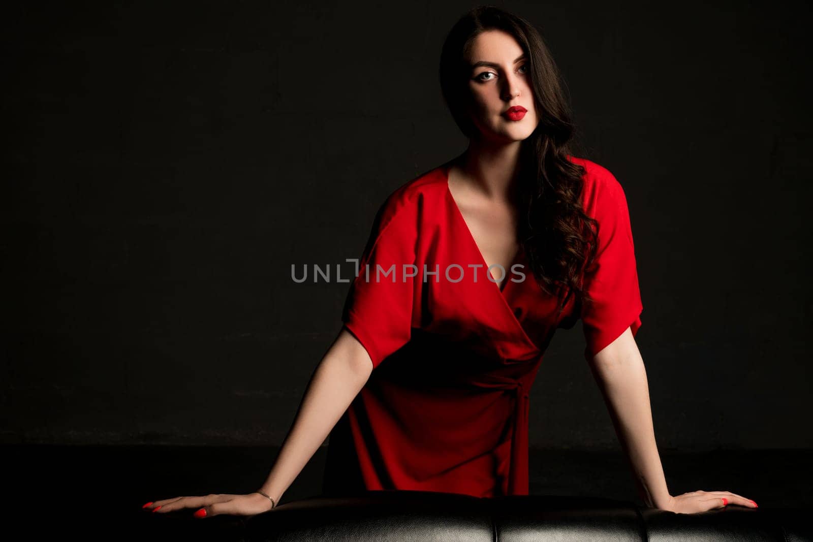 Sexy brunette in red dress leaning against black leather sofa shot in the dark room