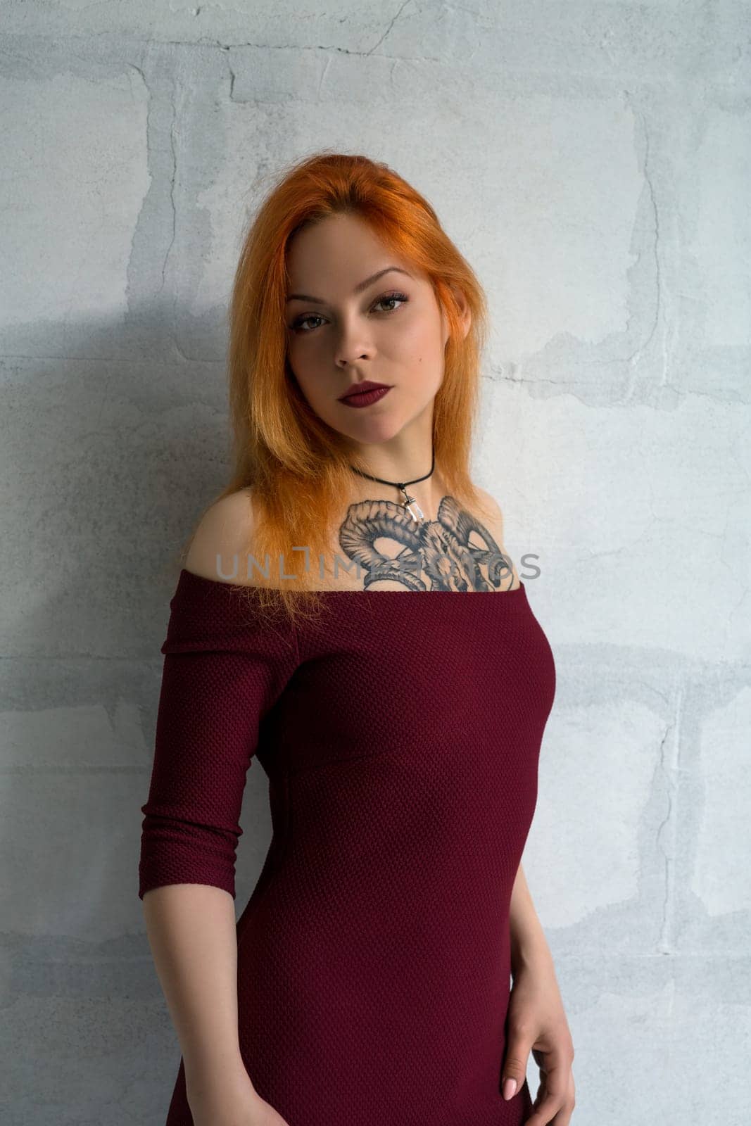 Hot red-haired girl with a tattoo wearing burgundy dress shot against wall