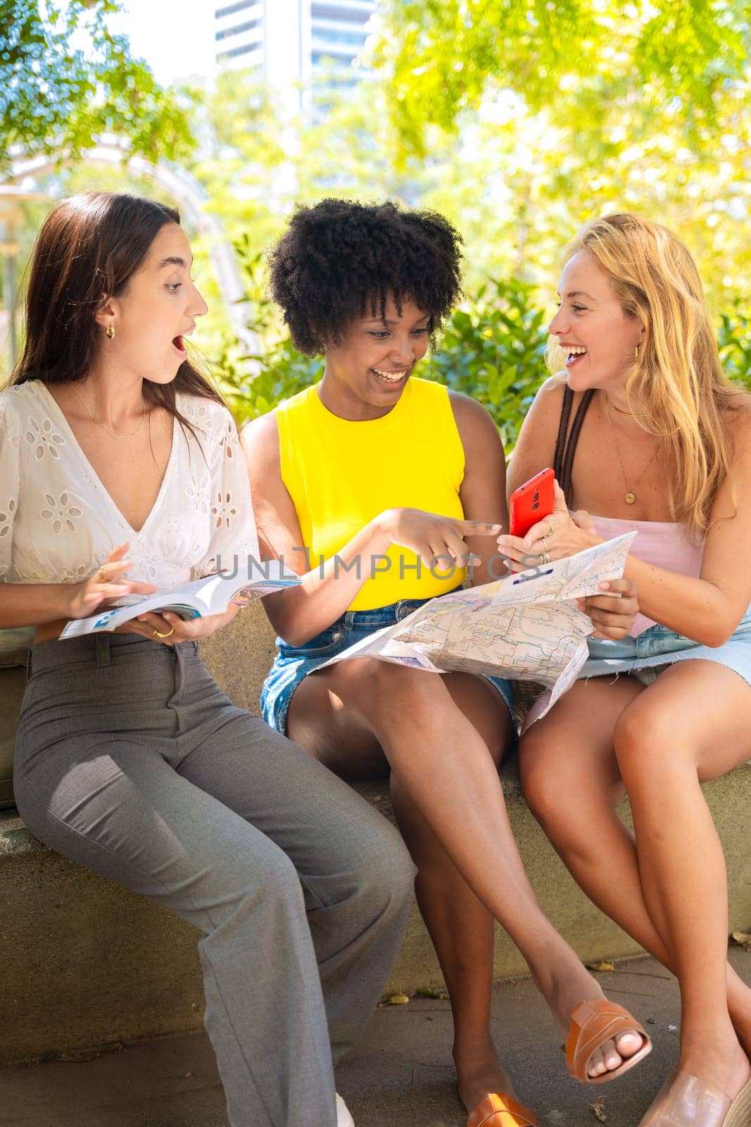 Three happy young women stand outdoors, looking at maps and a travel guide. They smile while planning their trip, enjoying a fun adventure in a sunny urban setting with trees and buildings.