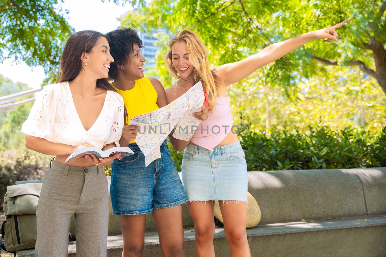 Three cheerful young women stand outdoors, holding maps and a travel guide while pointing at something. They smile excitedly, enjoying their adventure in a sunny urban setting with trees and buildings