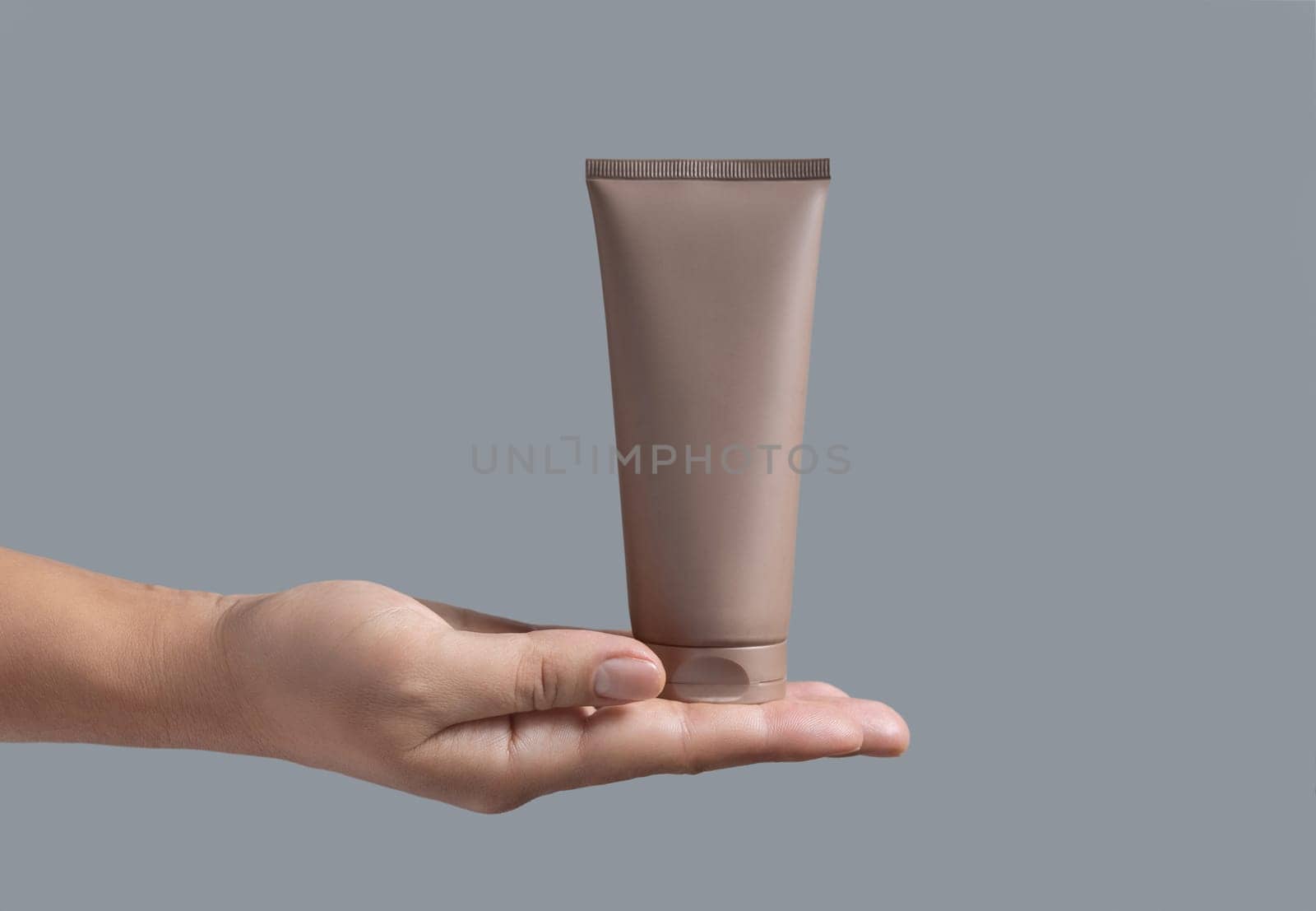 Female hand holding beige squeeze cream tube against grey close up, cosmetic package mockup, copy space. Skincare beauty product with minimal composition