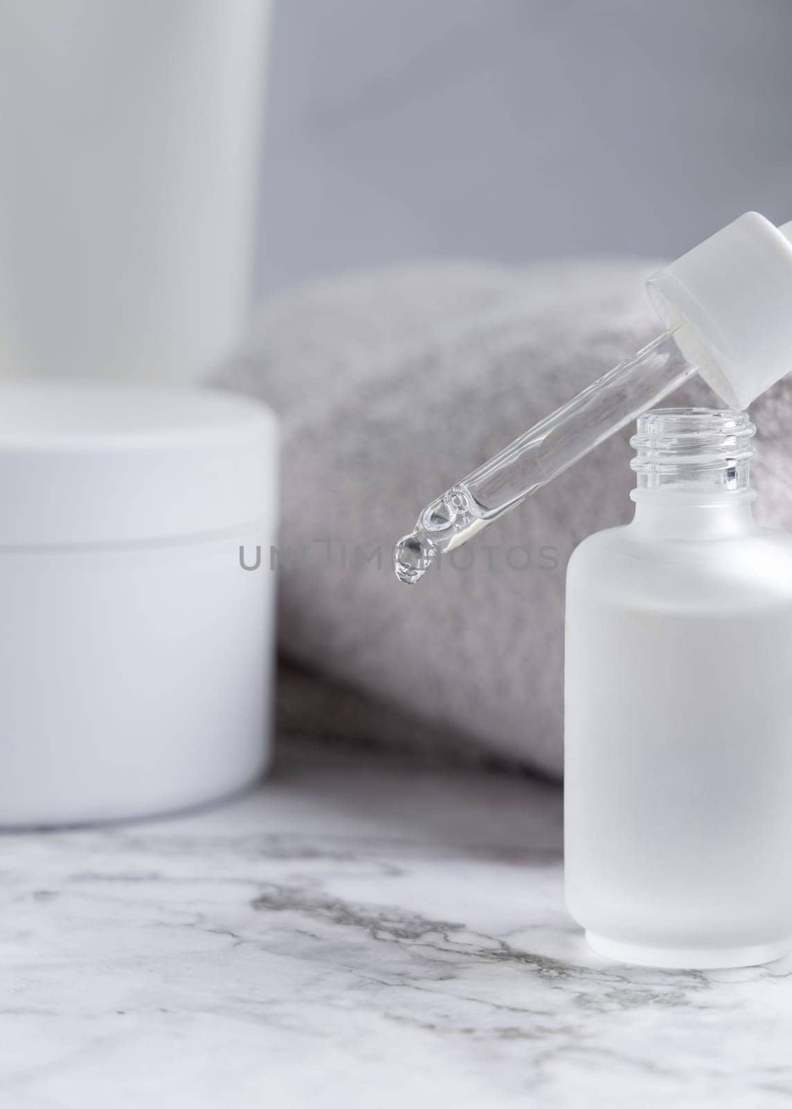 Drop falling from pipette set on a trasparent glass dropper bottle on white marble near cosmetic bottles and jars close up, packaging mockup. Natural serum or essence