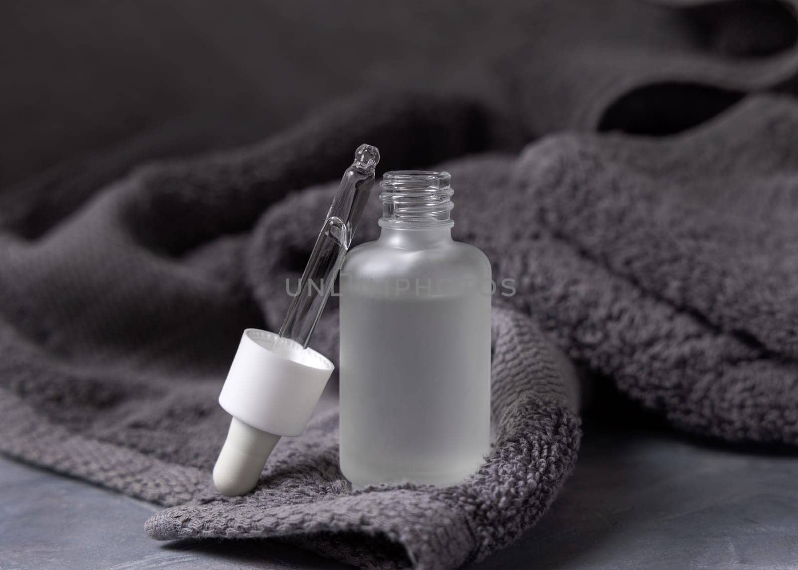 Pipette set on opened clear glass dropper bottle near dark grey bath towel closeup, brand packaging mockup. Natural handmade cosmetic product for facecare and spa procedures - serum or essence