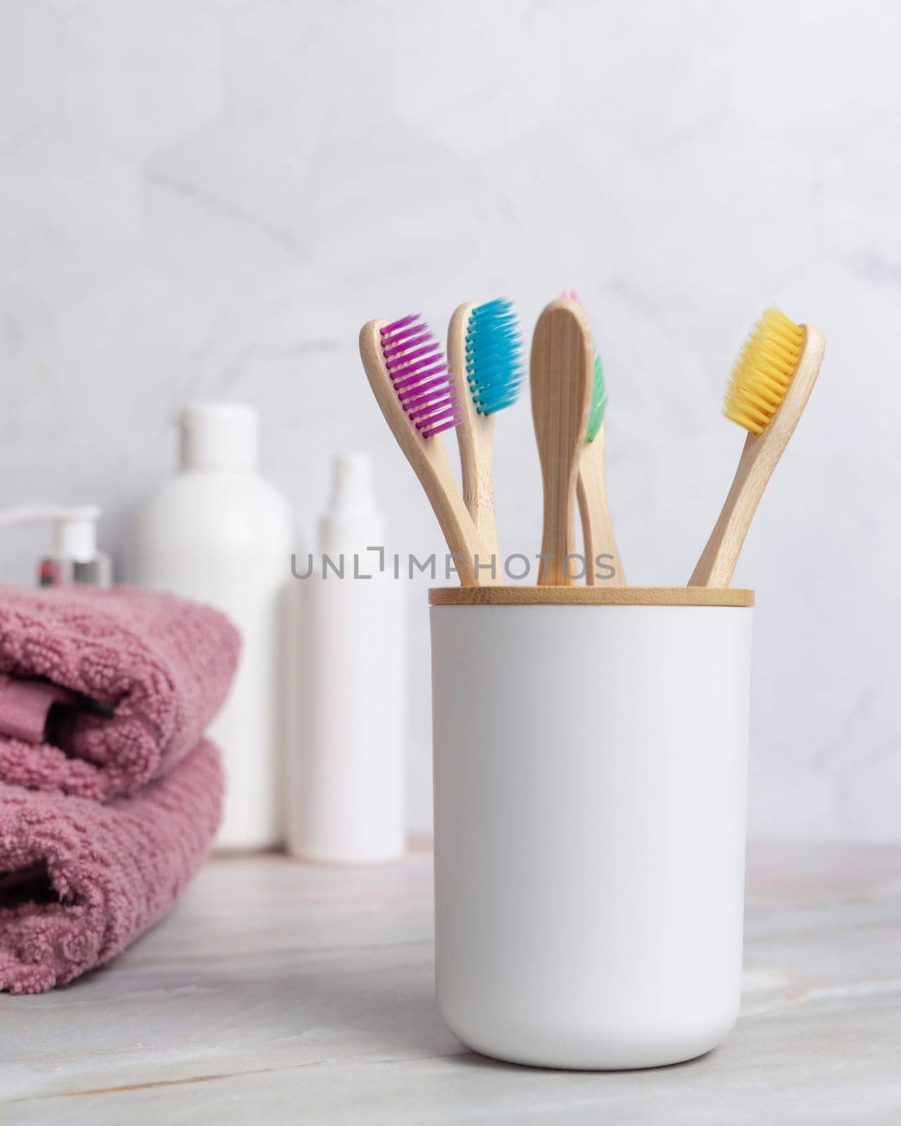 Eco-friendly colorful bamboo toothbrushes in holder cup in a white in bathroom against natural cosmetics and towel, mockup. Personal care and hygiene products