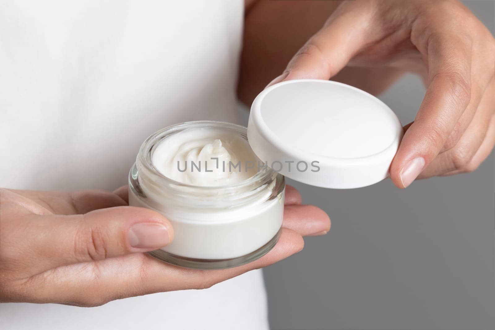 Woman opening cream jar against white shirt and grey background, fingers with natural manicure, cosmetic mockup. Concept of organic cosmetics for body or face care