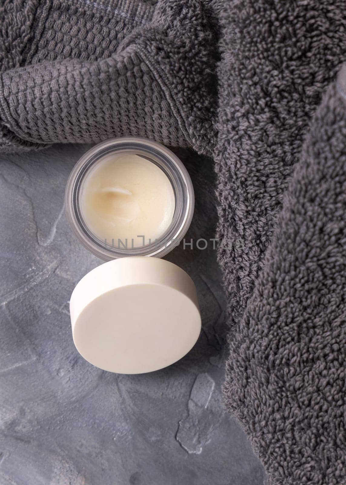 Opened lip balm jar with blank lid near dark grey folded towel top view, cosmetic packaging mockup, copy space. Lipcare beauty product for mens everyday personal care