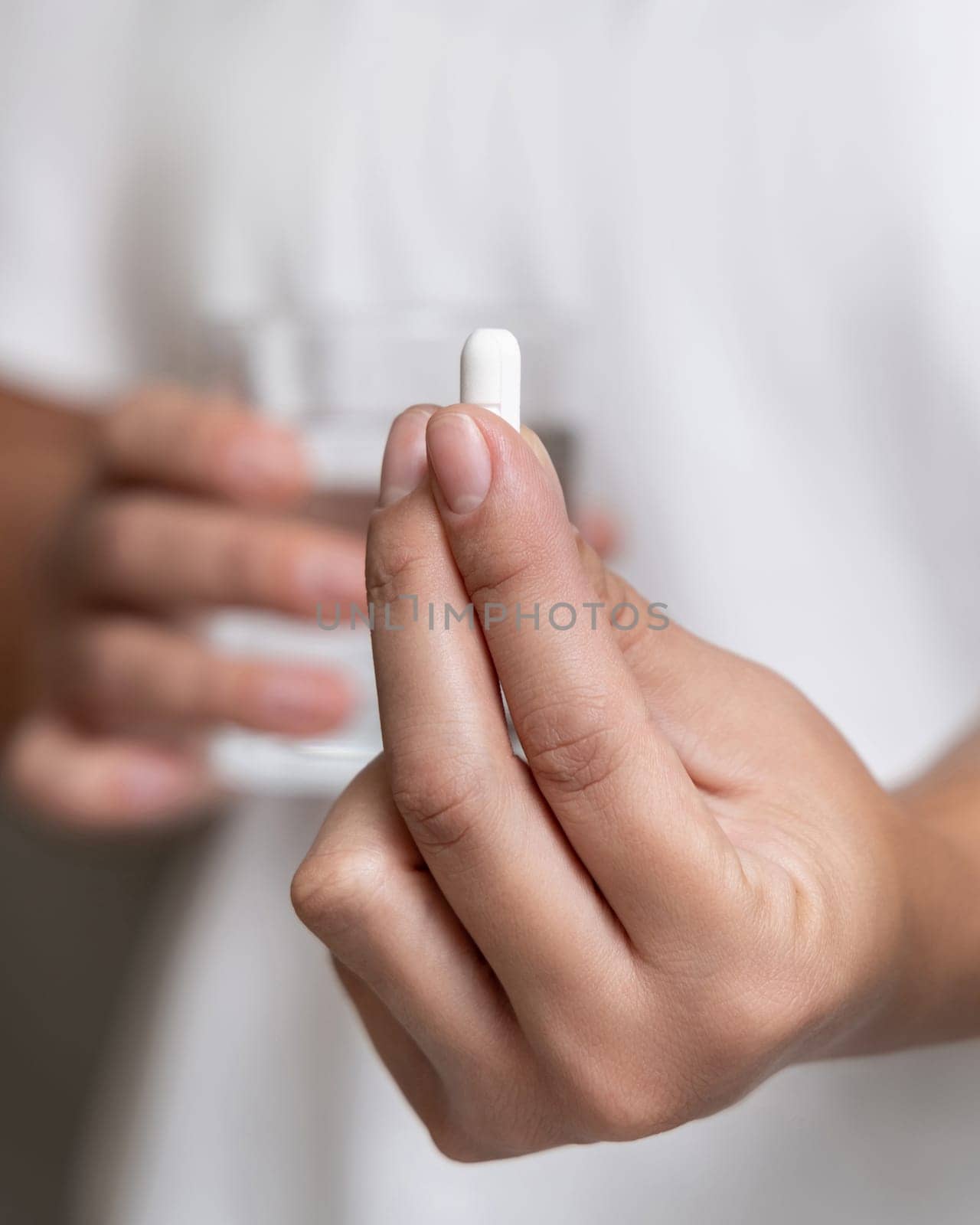 Woman in white t-shirt holding white pill in fingers and water glass in hand closeup. Female or male taking dietary supplement, daily vitamins. Healthcare concept