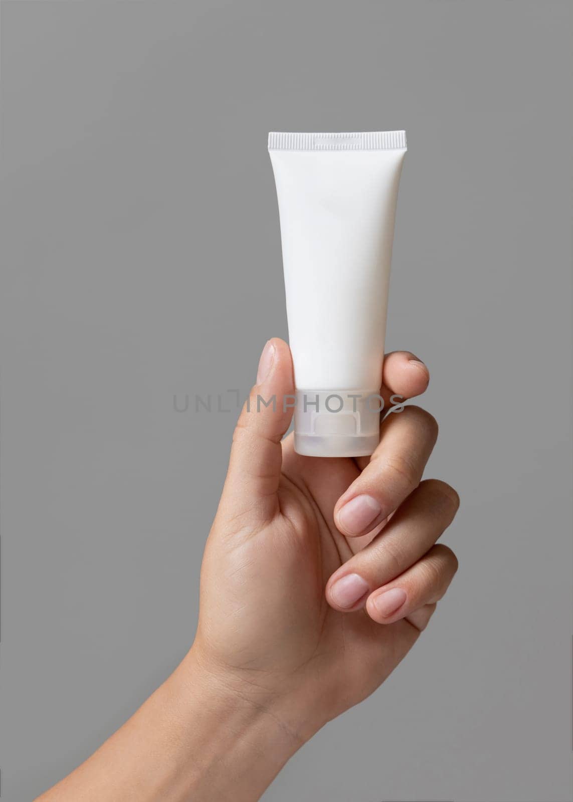 Female hand holding white squeeze cream tube against grey close up, cosmetic package mockup, copy space. Skincare beauty product with minimal composition