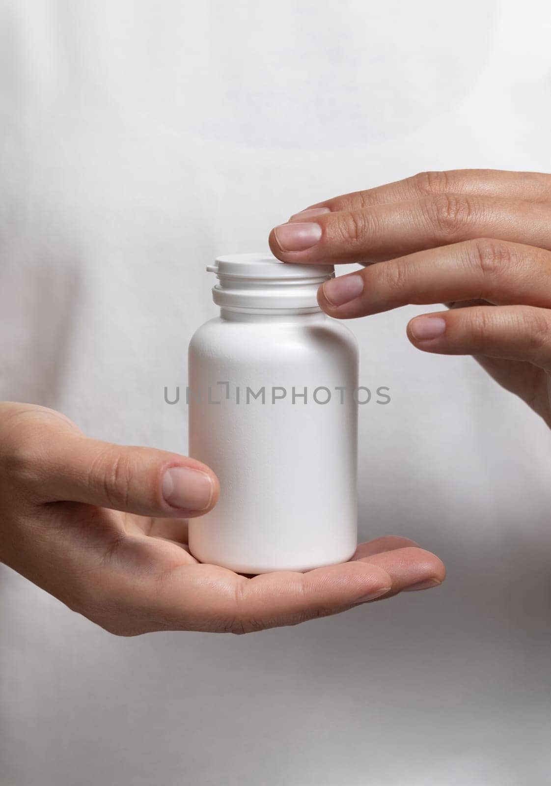 Closeup of woman in white t-shirt holding a white plastic medical pill bottle. Packaging mockup for herbal supplement. Female hands with natural manicure