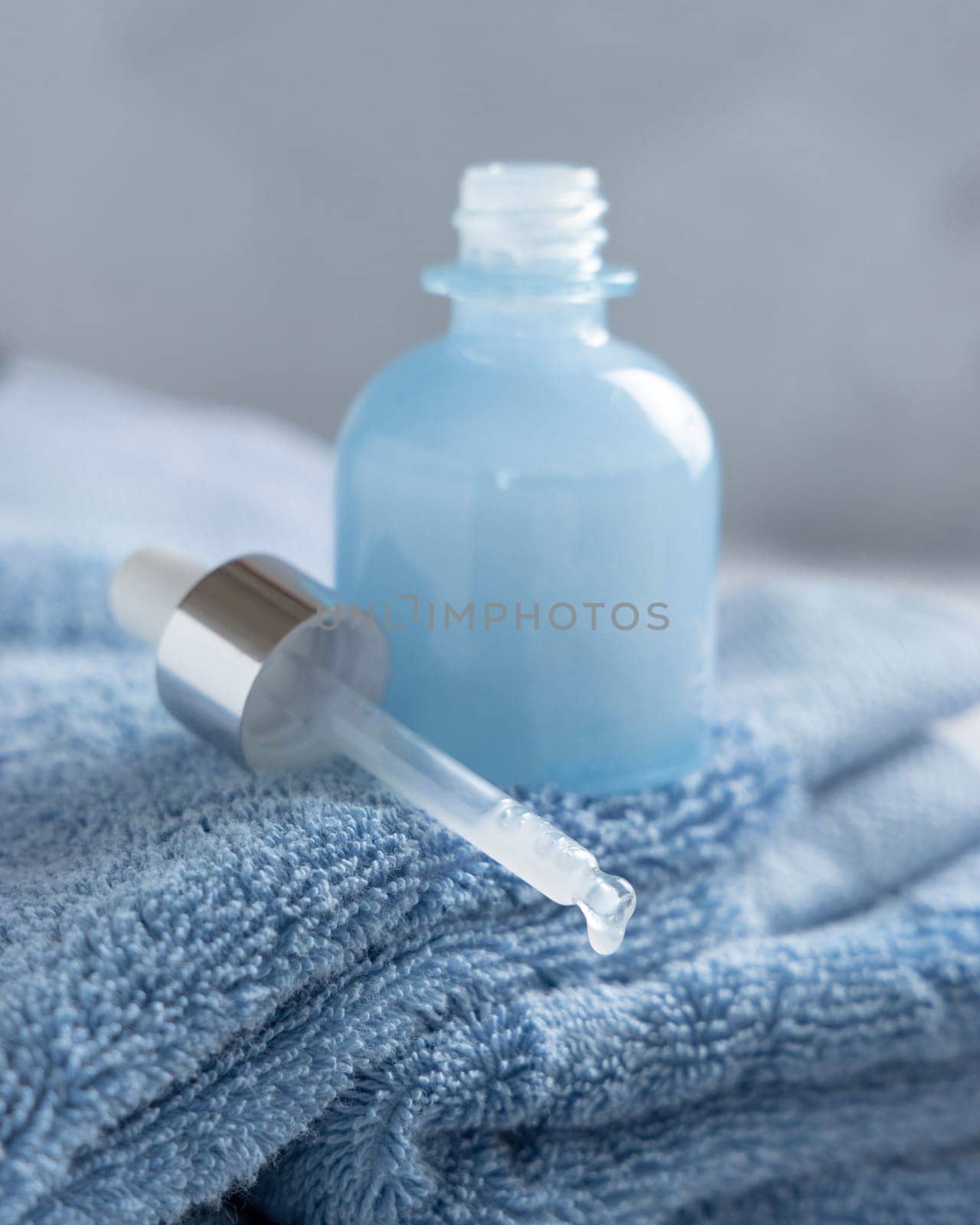 Drop falling from pipette put near opened blue glass dropper bottle on blue towel against grey wall close up, mockup. Natural serum or essence