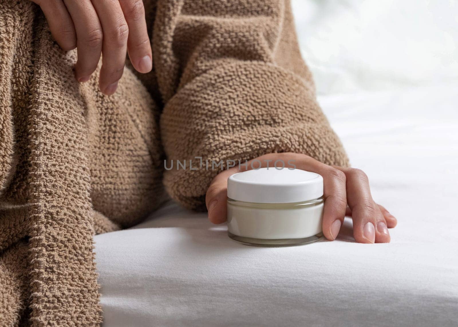 Woman in brown bathrobe puts white opened cream jar on white bedsheet closeup. Bodycare concept. Organic cosmetics for body and face care, beauty and spa procedures