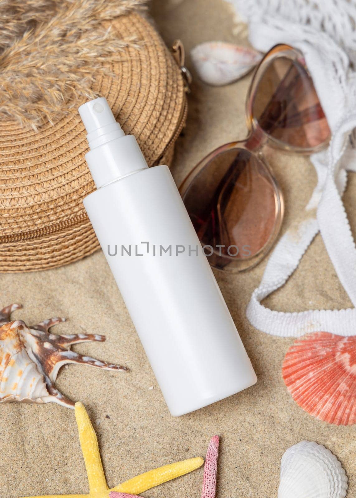 White blank spray bottle, sunglasses, towel, starfishes and shells on a sandy beach, top view. Sunscreen cosmetics mockup. Summer vacation and spf uv-protecting skin care concept