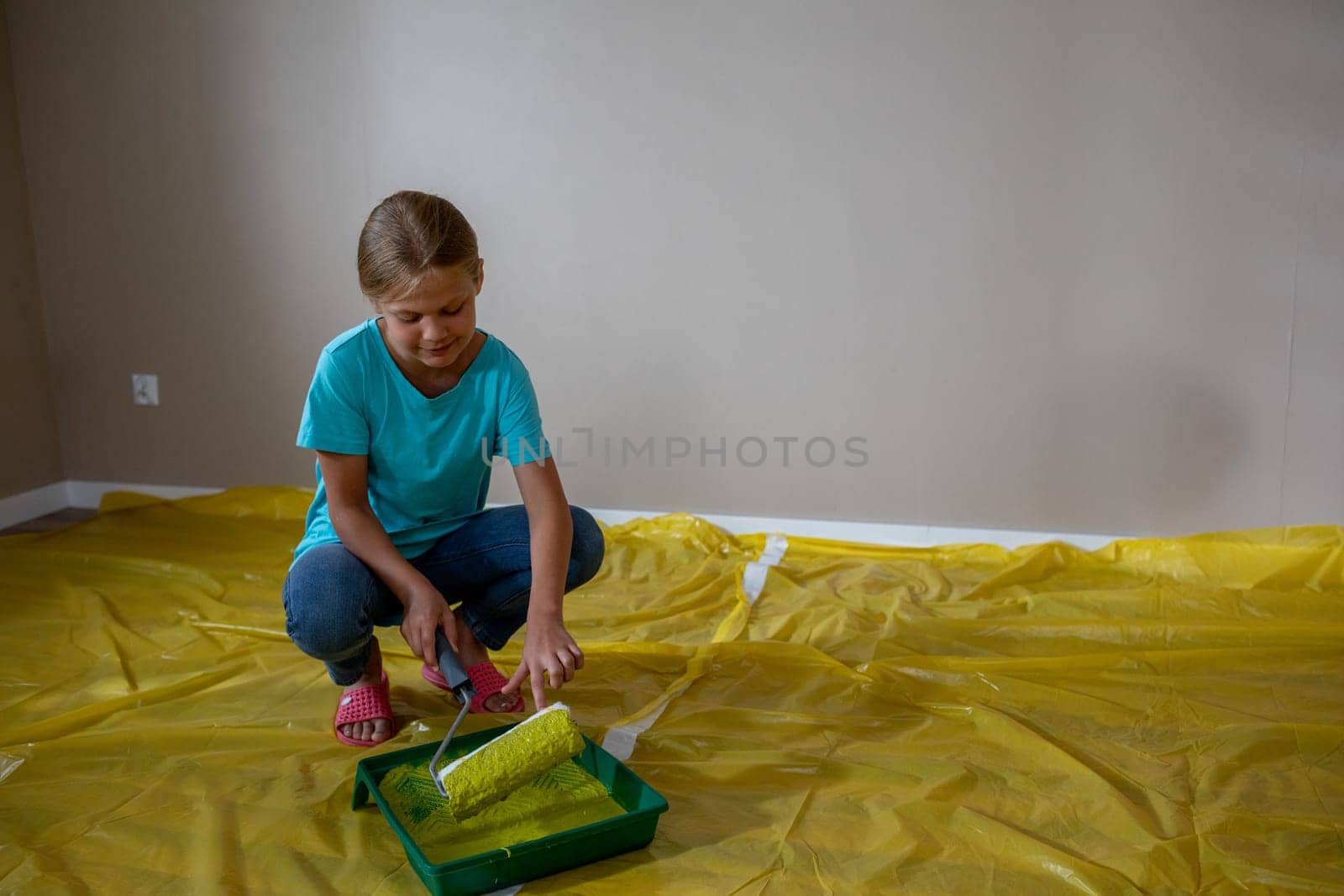 A child excitedly gets ready to paint a whole room using a roller while standing on a bright yellow plastic sheet