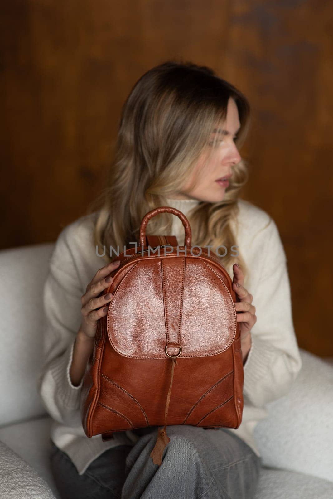 beautiful girl in gray pants and beige sweater posing with a small brown leather backpack.