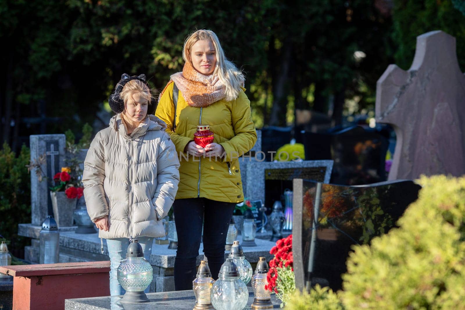 A mother and her young child visit a peaceful cemetery, honoring family with candles and vibrant flowers
