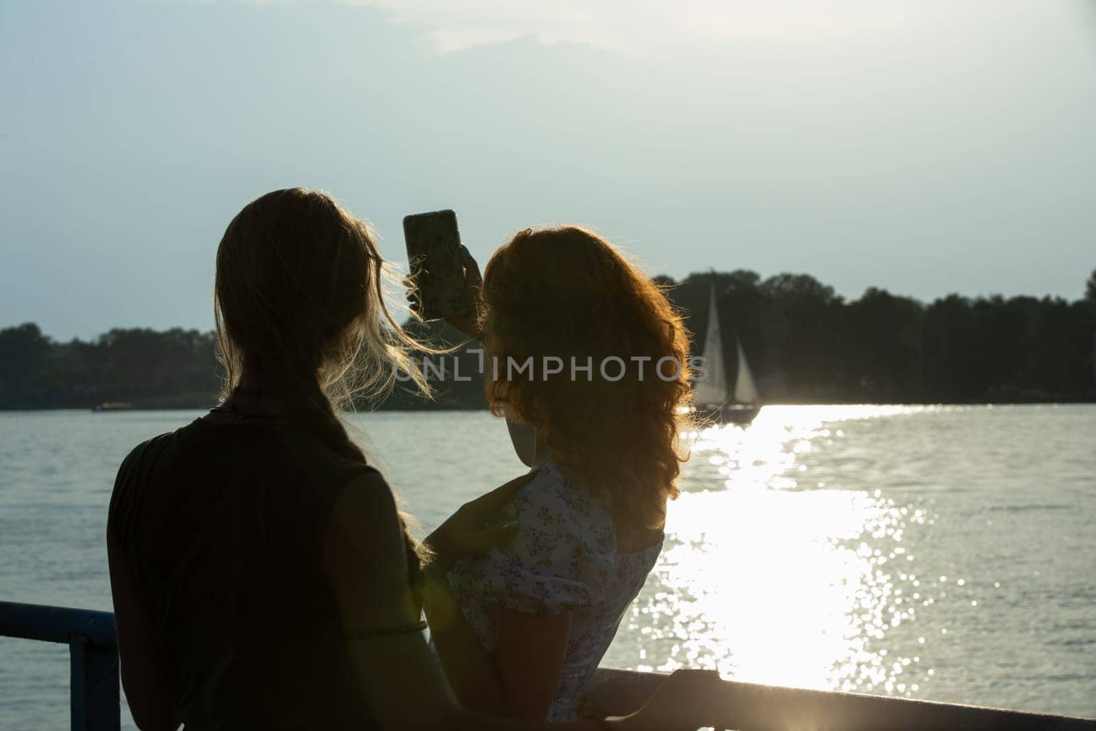 Two friends happily take a sunset selfie by the peaceful water, celebrating their friendship and adventure