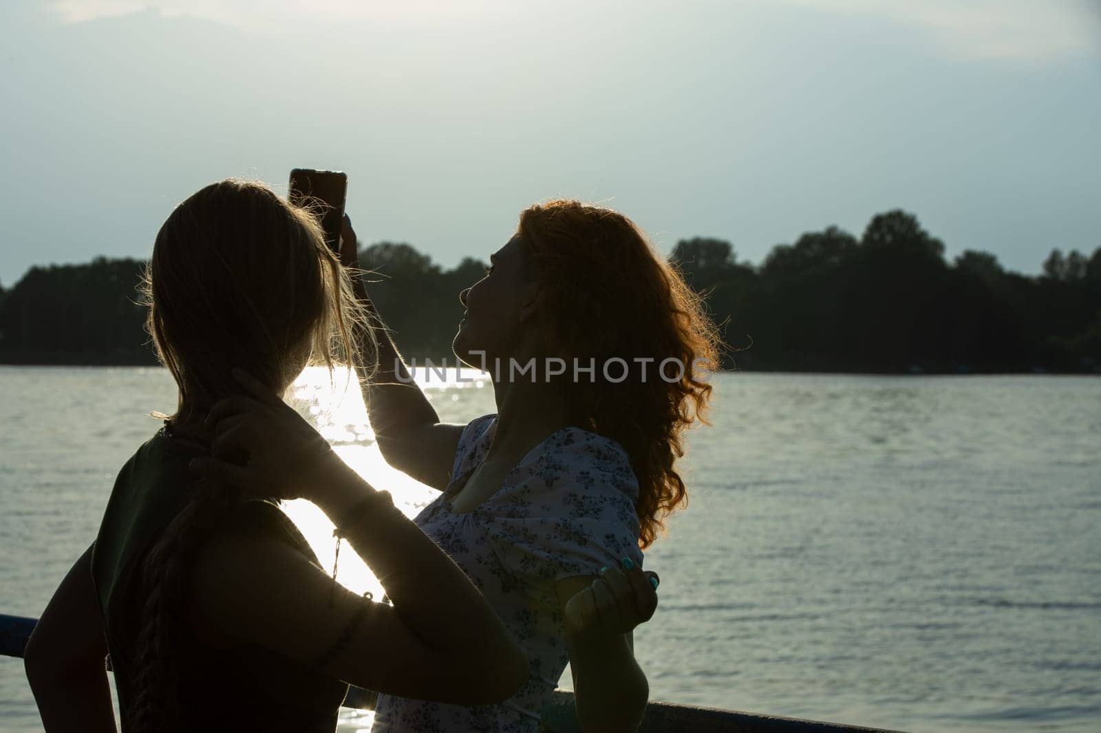 In this peaceful scene, two friends joyfully take selfies by the shimmering water during a beautiful sunset