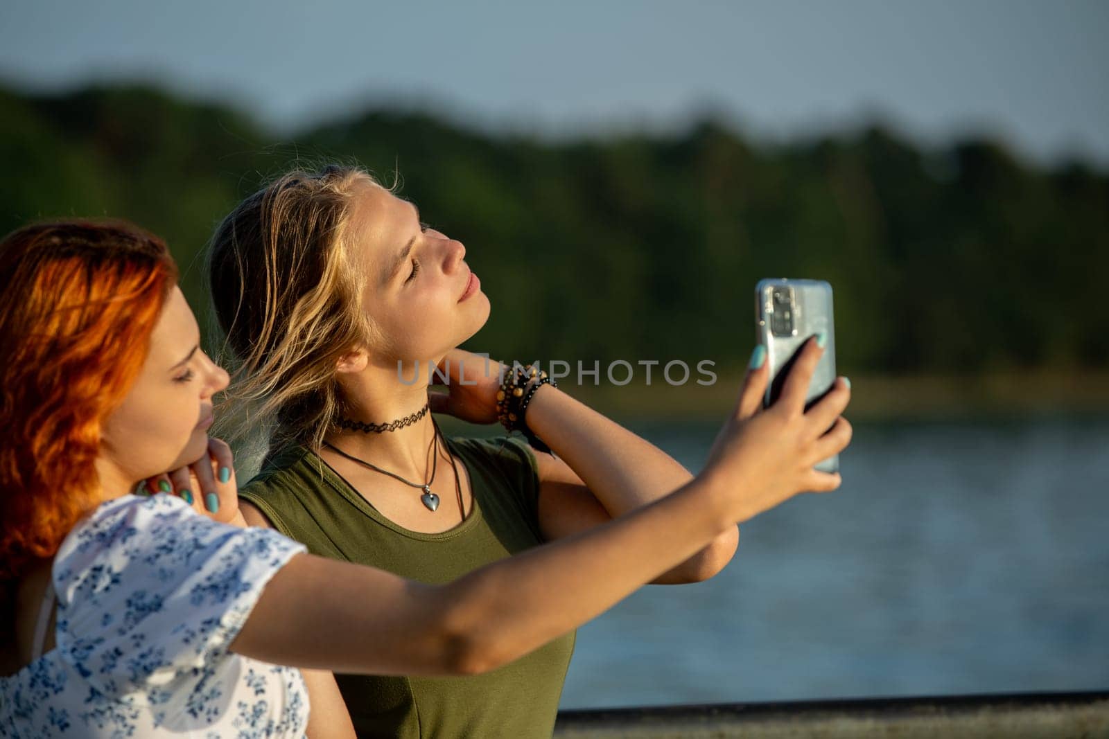 Two young women happily take a selfie by the shimmering water, capturing a fun and joyful moment in the bright sun