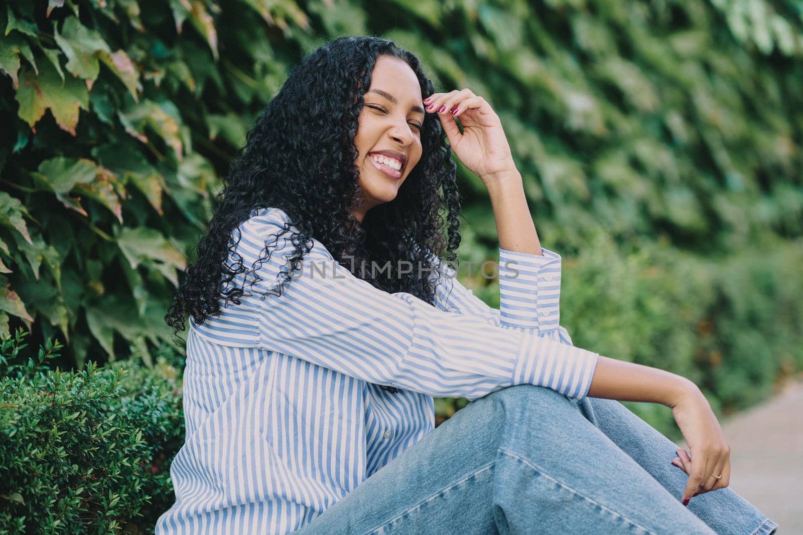 A cheerful and vibrant young woman is seated comfortably against a lush green backdrop, wearing a stylish striped shirt and smiling joyfully, truly embodying happiness and positivity in her presence