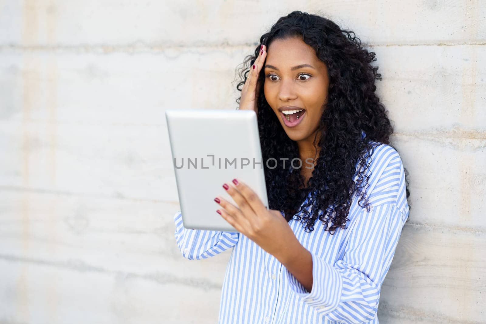 A young woman with beautifully curly hair radiates surprise and beams with joy while enthusiastically using a tablet, showcasing a lively moment filled with genuine engagement and deep emotion