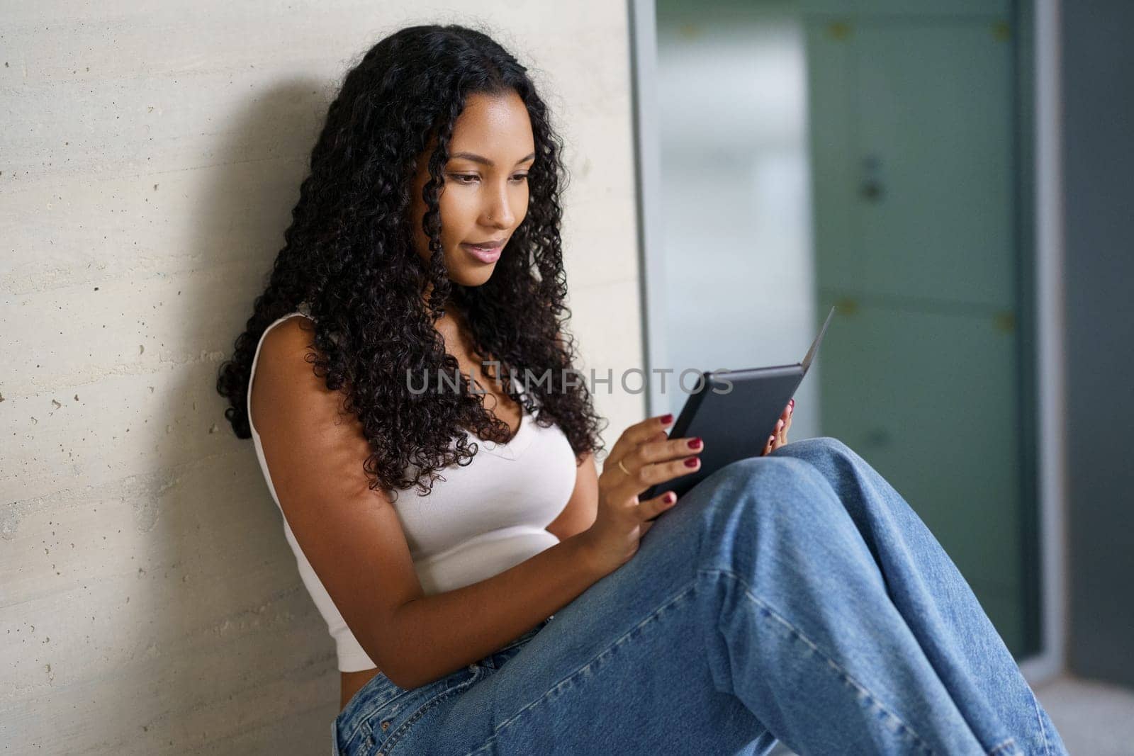 A young woman with beautifully styled curly hair sits comfortably against a chic wall, fully engrossed in her tablet, enjoying a joyful and peaceful moment of leisure and modern technology