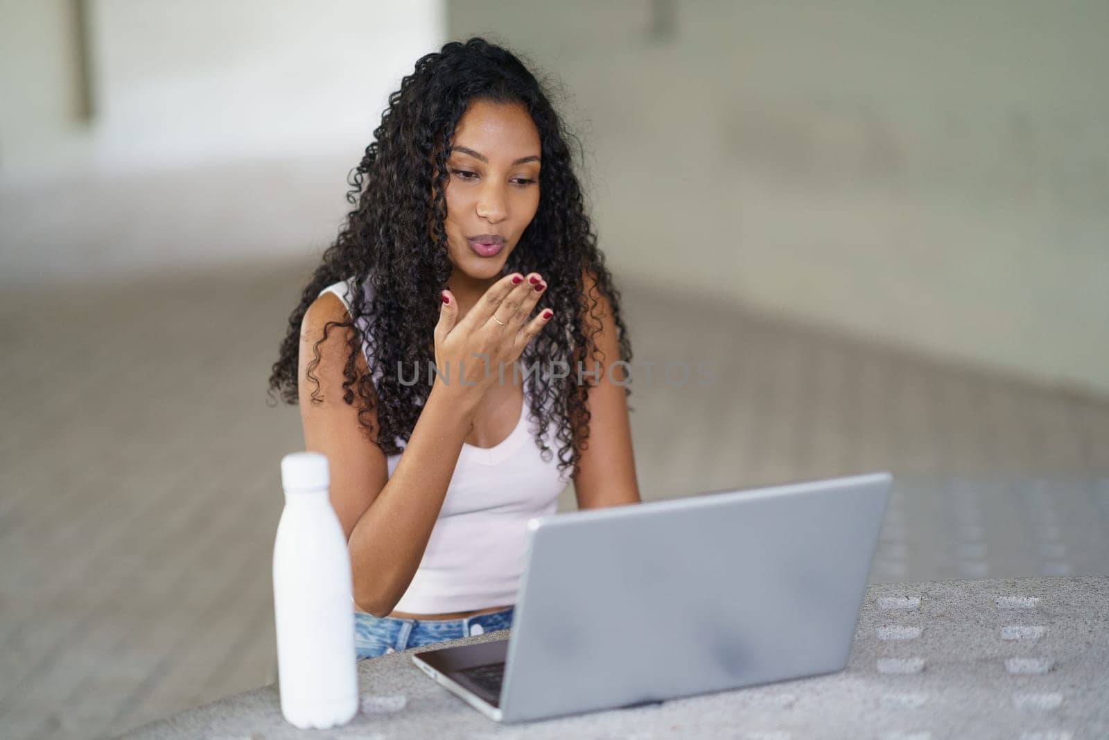 A young woman with beautiful curly hair is engaged in an important video call on her laptop, focused and surrounded by a neat, minimalist background that enhances her modern workspace