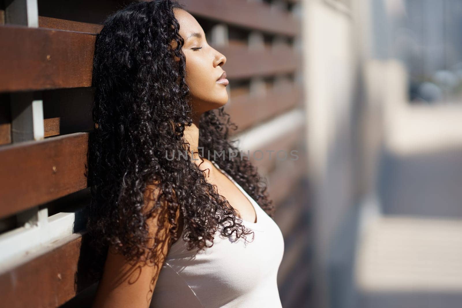 A serene and peaceful woman with long, flowing curly hair is enjoying the warm sunlight, comfortably leaning against a rustic wooden wall, which evokes a strong sense of calmness and relaxation