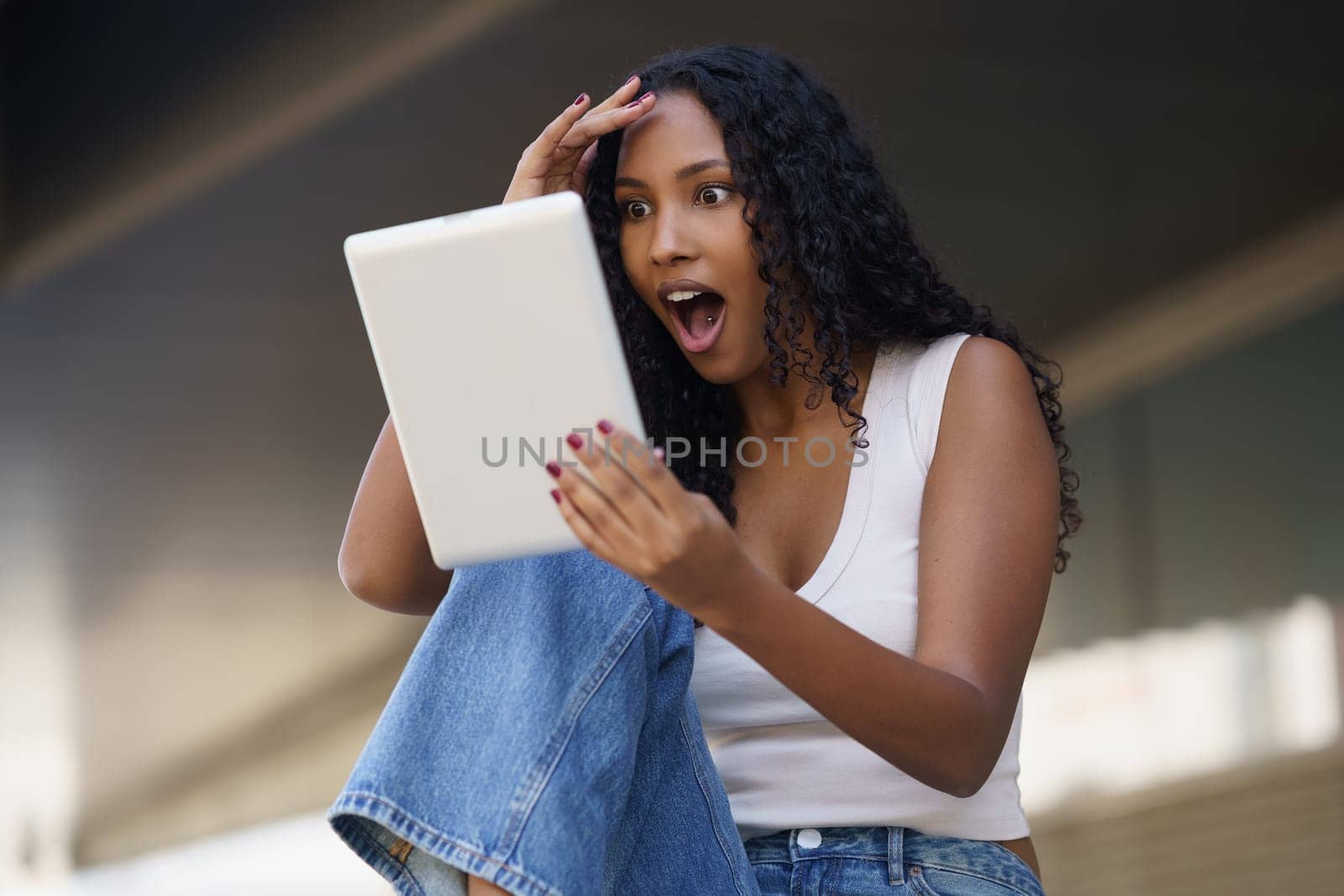 A young woman is using her tablet in an outdoor setting, and as she looks at the screen, her face lights up with genuine surprise and excitement, showcasing her deep engagement and enthusiasm