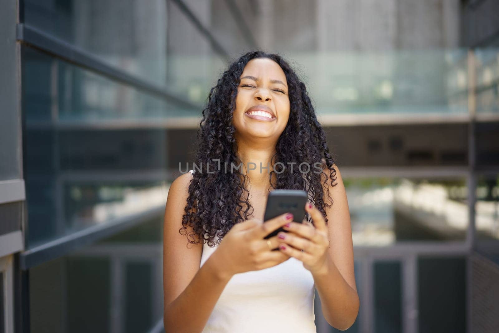 A cheerful and vibrant young woman holds her smartphone up while smiling widely, fully enjoying a lovely sunny day in a lively urban setting filled with energy and excitement all around her