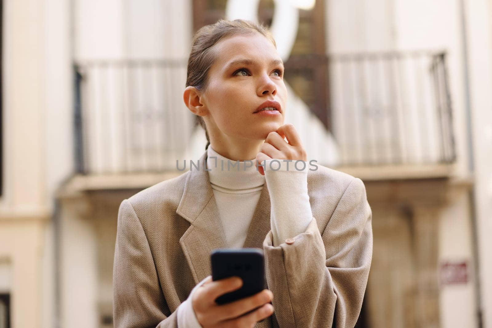 In a charming urban setting, a young woman in an elegant coat and cozy turtleneck focuses on her smartphone, lost in thoughts while enjoying her beautiful surroundings with a sense of peace