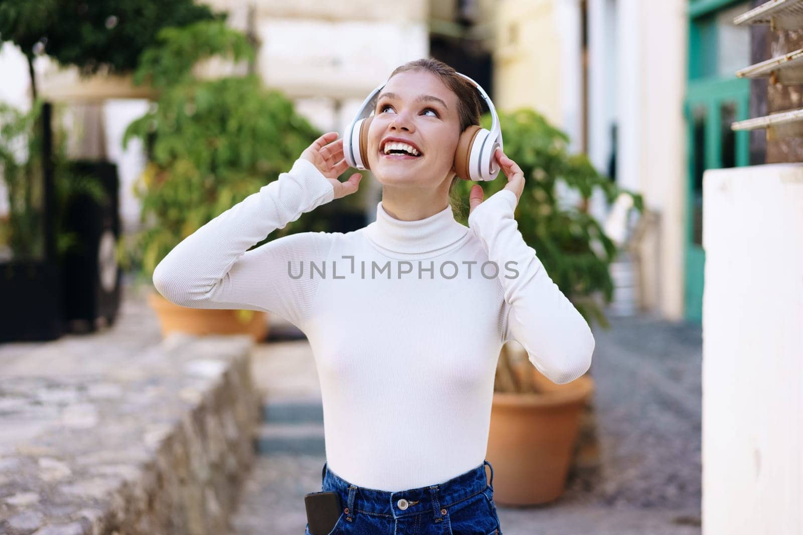 In an urban setting, a cheerful young woman with stylish headphones joyfully listens to music outdoors, embodying happiness, relaxation, and the exhilarating freedom that music brings