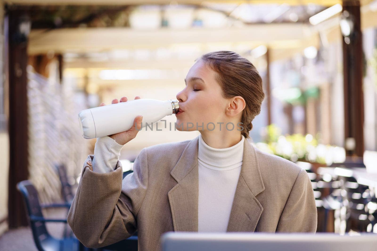 In a trendy outdoor cafe, a stylish woman confidently sips from a chic water bottle, exuding a sense of relaxation as she takes in and enjoys her beautiful surroundings on a lovely day