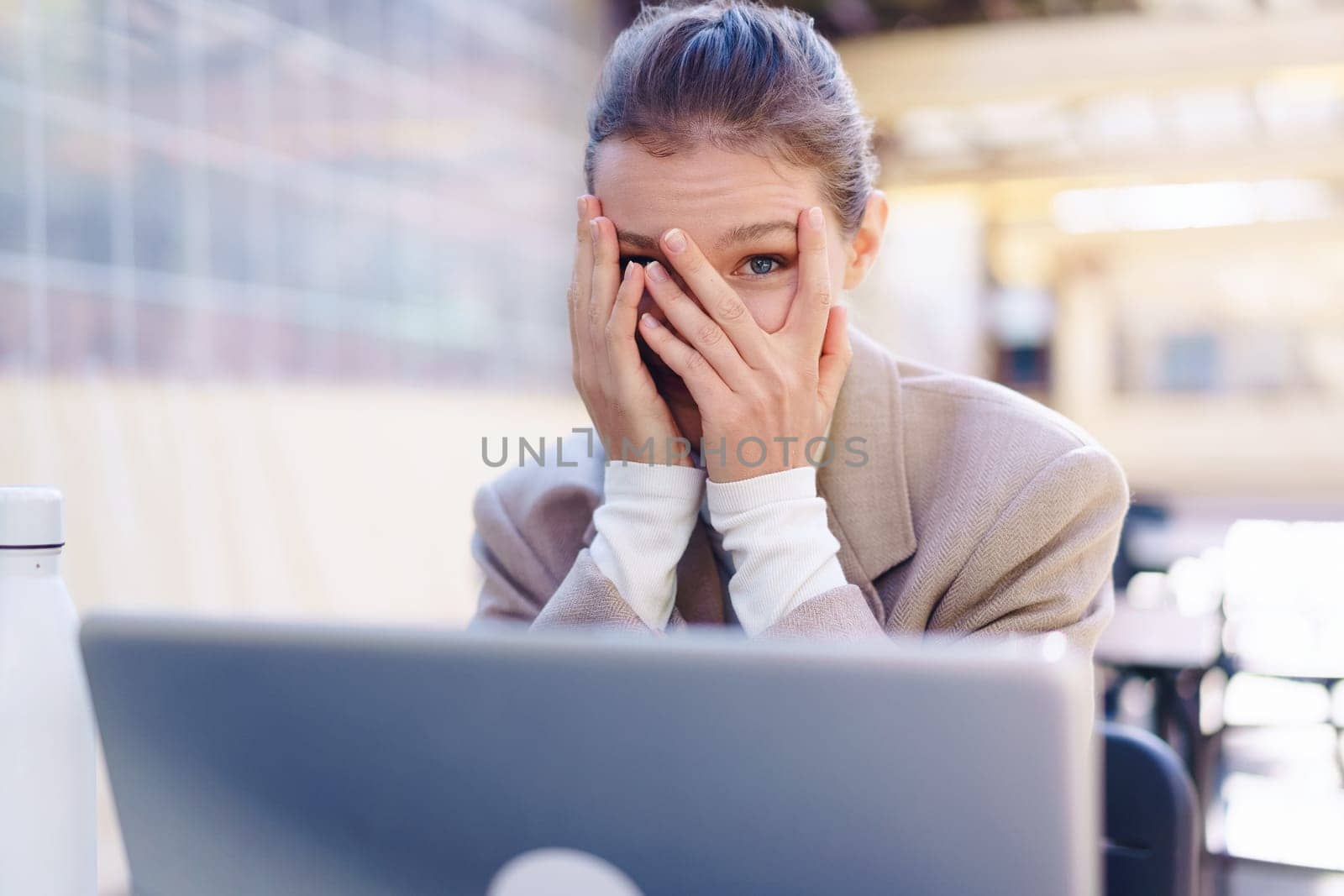 A stressed businesswoman sits at a laptop, covering her face with her hands, showing intense pressure and emotional exhaustion from her professional struggles and productivity demands