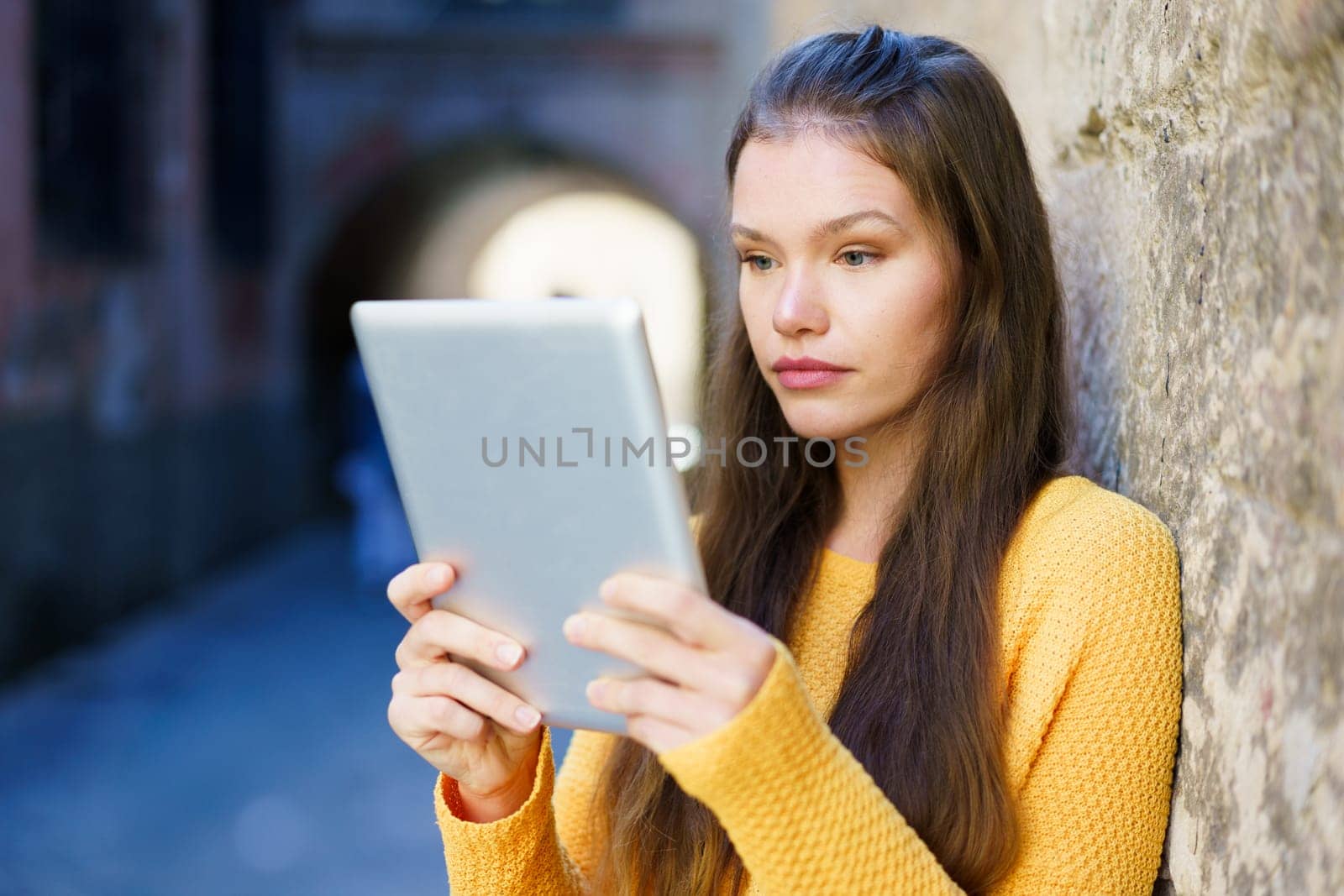 A young woman in a bright yellow sweater is intently using her tablet against a weathered stone wall in a lively urban setting, highlighting modern connectivity in our digital age