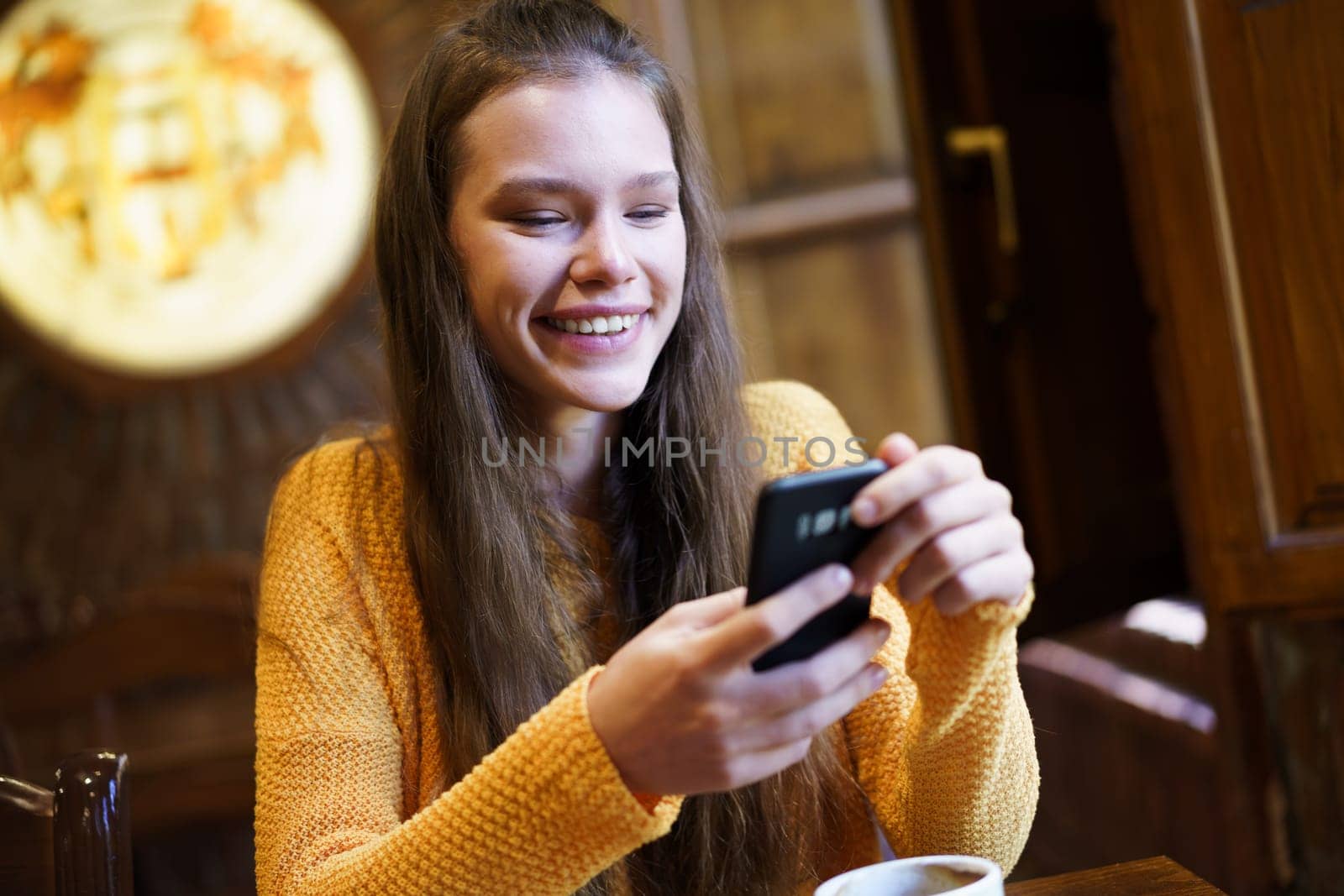 A young woman in a cozy caf smiles while texting on her smartphone. Warm lighting enhances the inviting atmosphere, perfect for social interactions