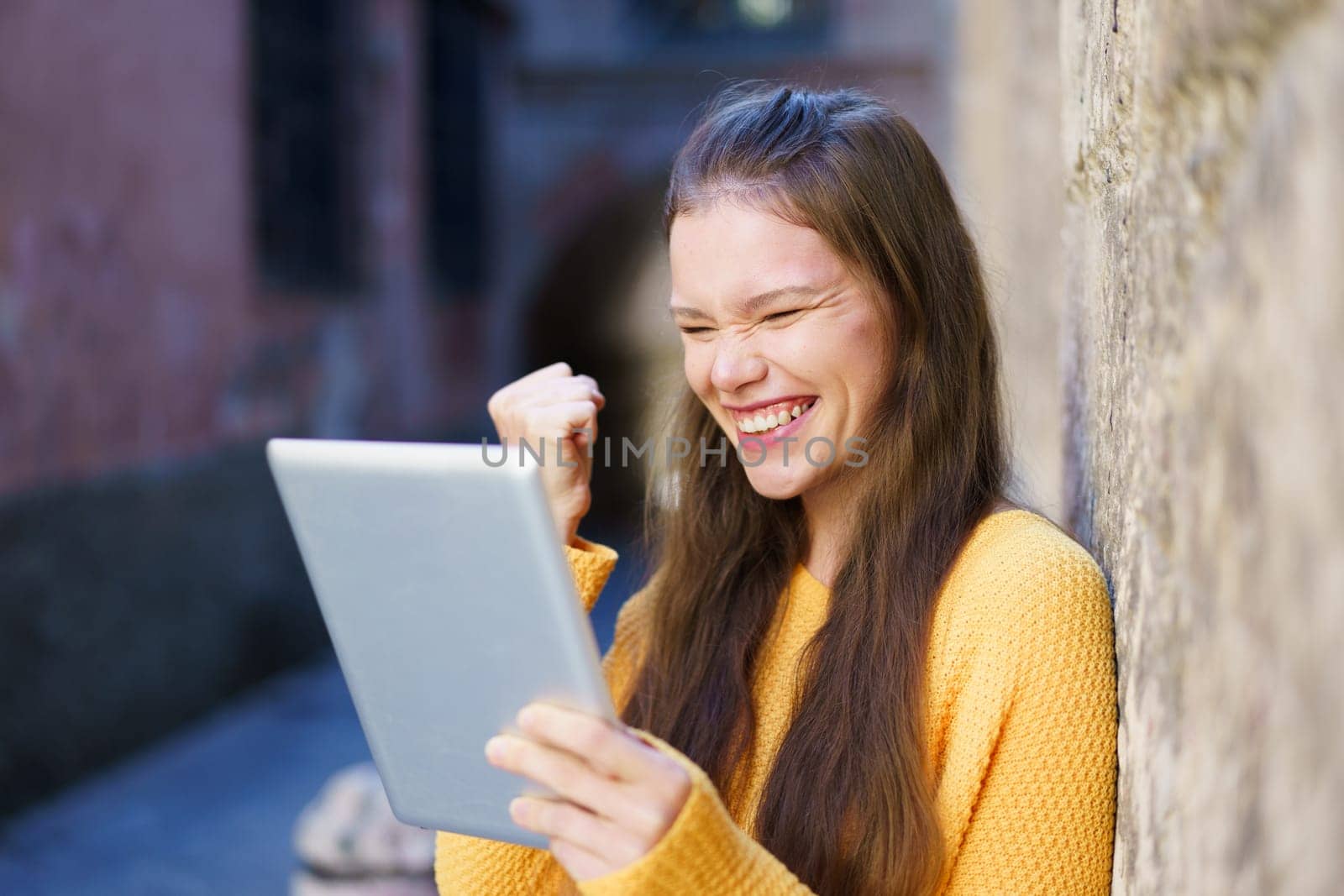 A young woman who is wearing a bright yellow sweater radiates joy as she smiles and celebrates, actively using a tablet, all set against a charming and historic backdrop that adds character