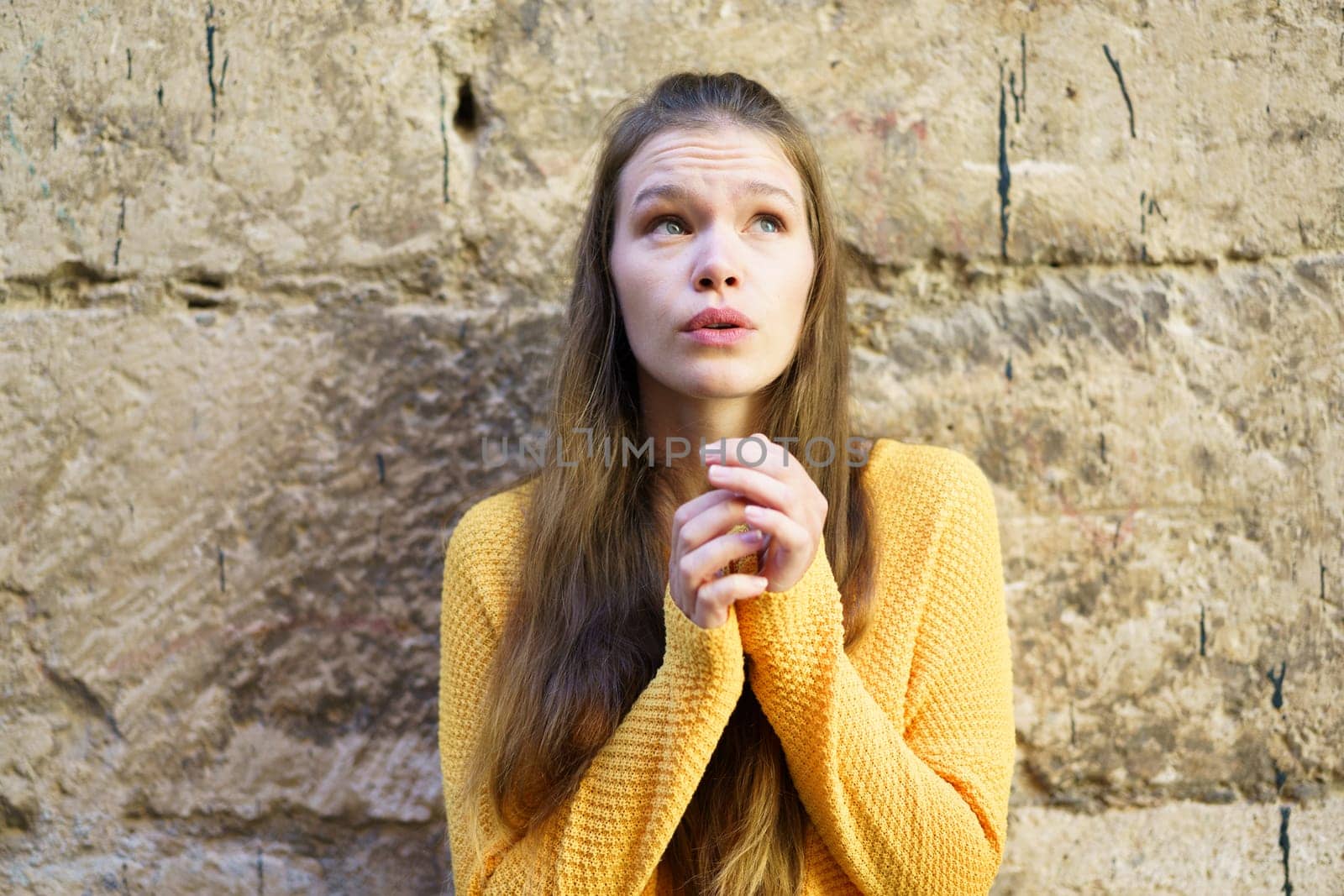 A young woman wearing a vibrant yellow sweater gazes thoughtfully upwards, her expression conveying a mixture of curiosity and hope, lost in her daydreams while leaning against a textured stone wall