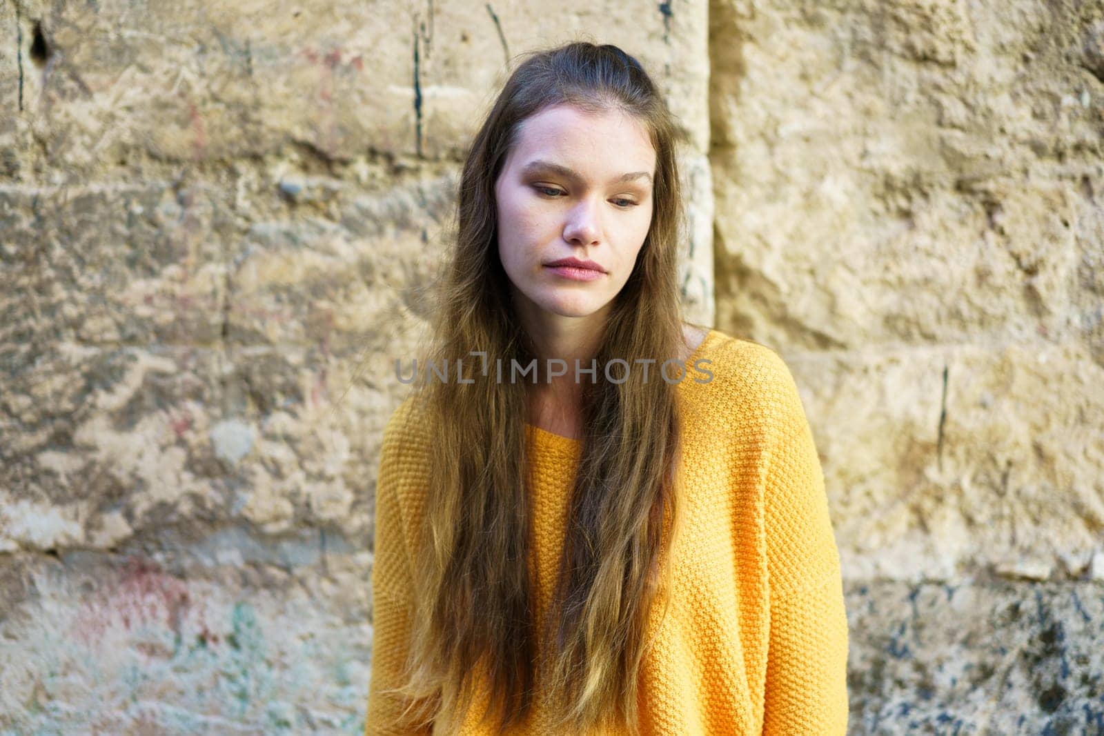 A young woman with long, flowing hair, dressed in a vibrant yellow sweater, stands in front of a textured stone wall, lost in deep contemplation, showcasing her beauty and calm demeanor